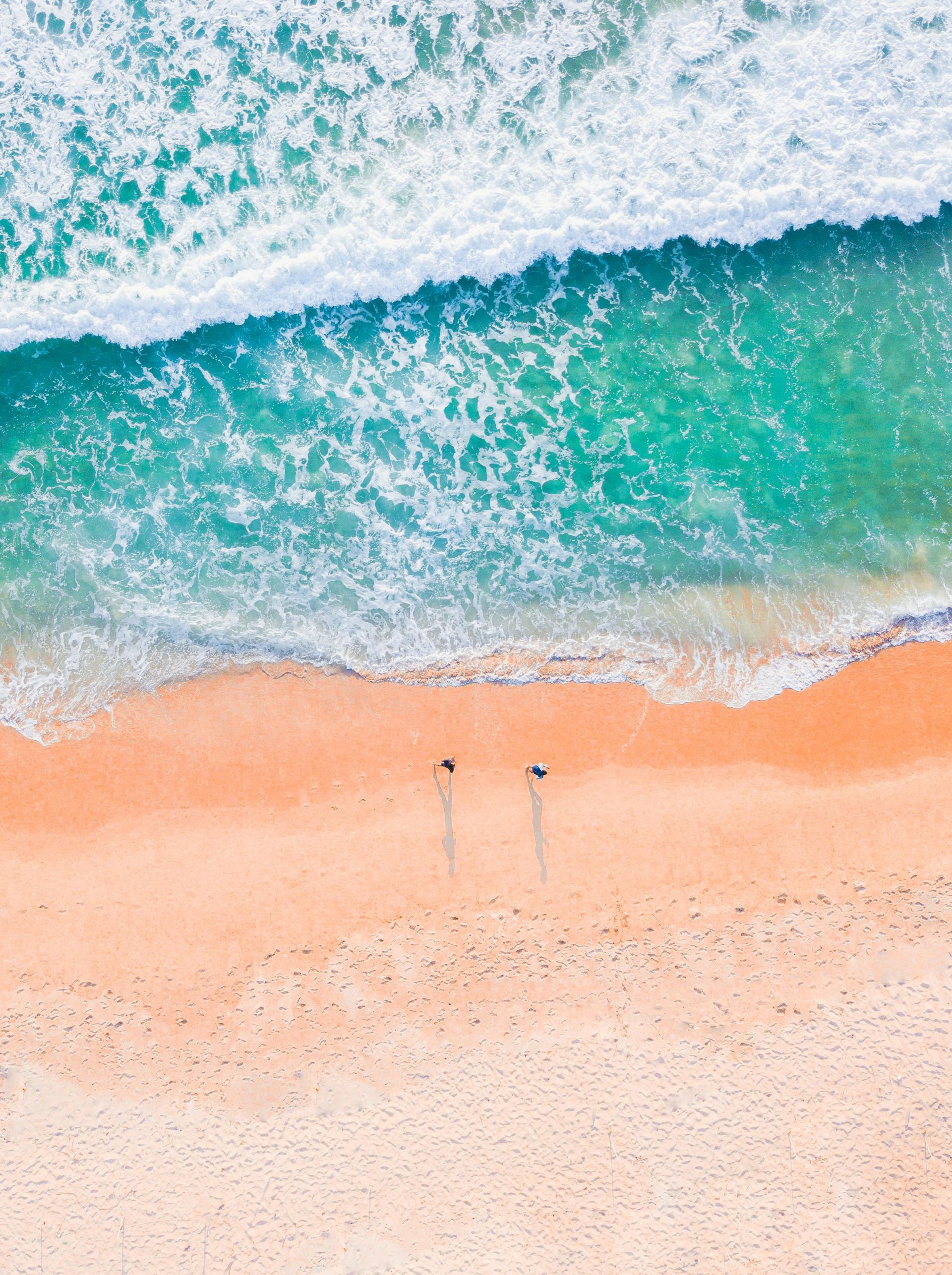 Aerial view of ocean waves crashing onto a sandy beach, two figures standing near the water.