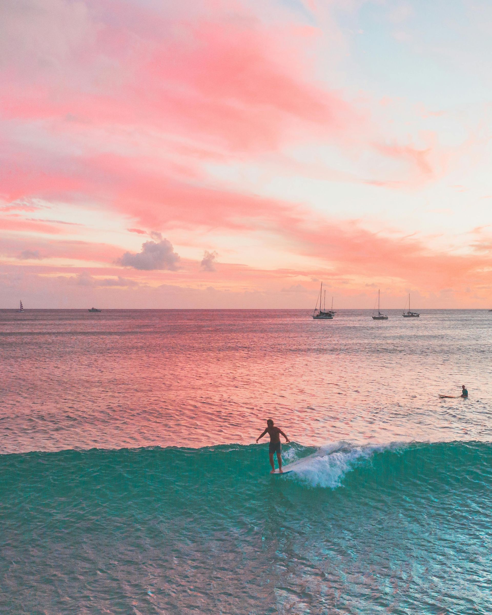 Surfer riding a wave at sunset, pink and orange sky over ocean, boats in the distance.