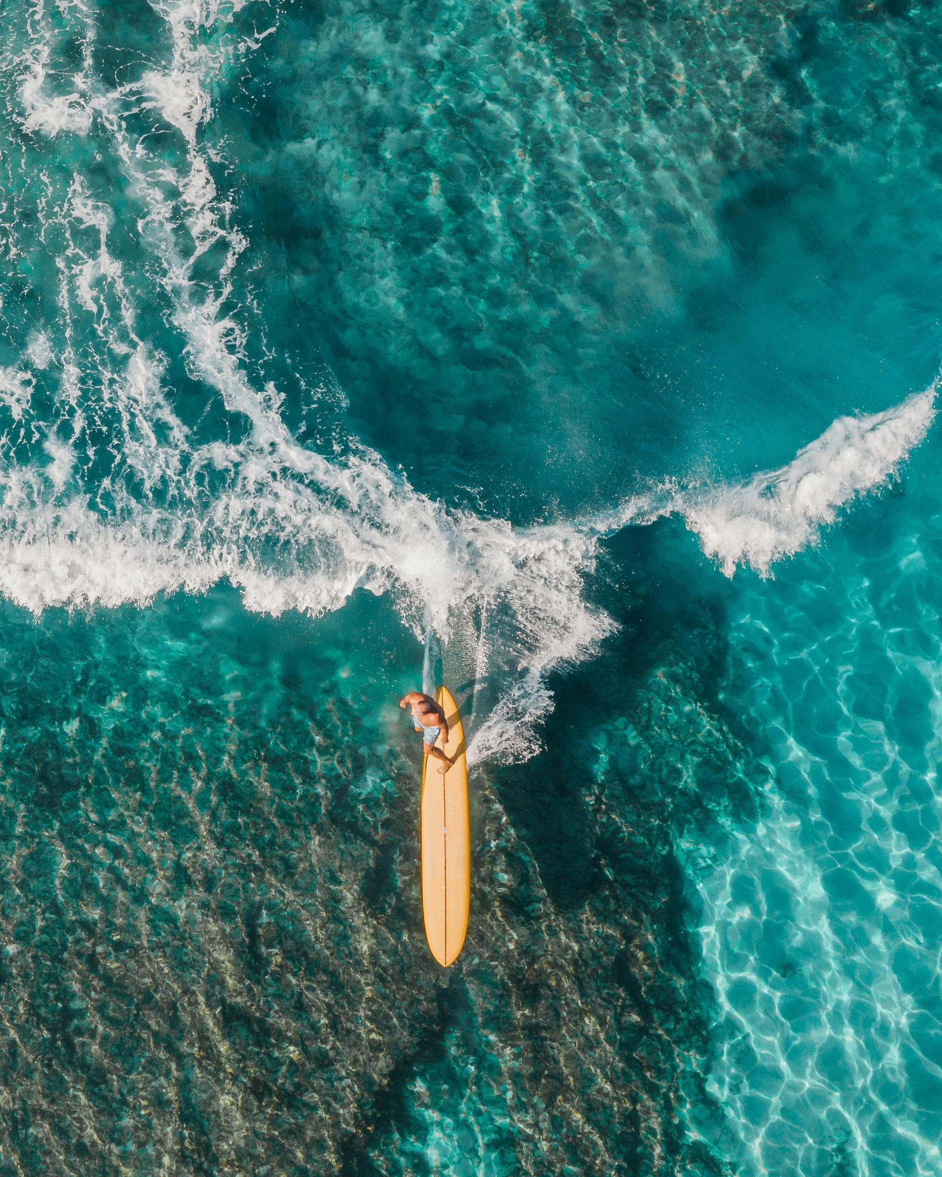 An aerial view of a person paddling on a long, yellow surfboard over vibrant turquoise water with a breaking wave.