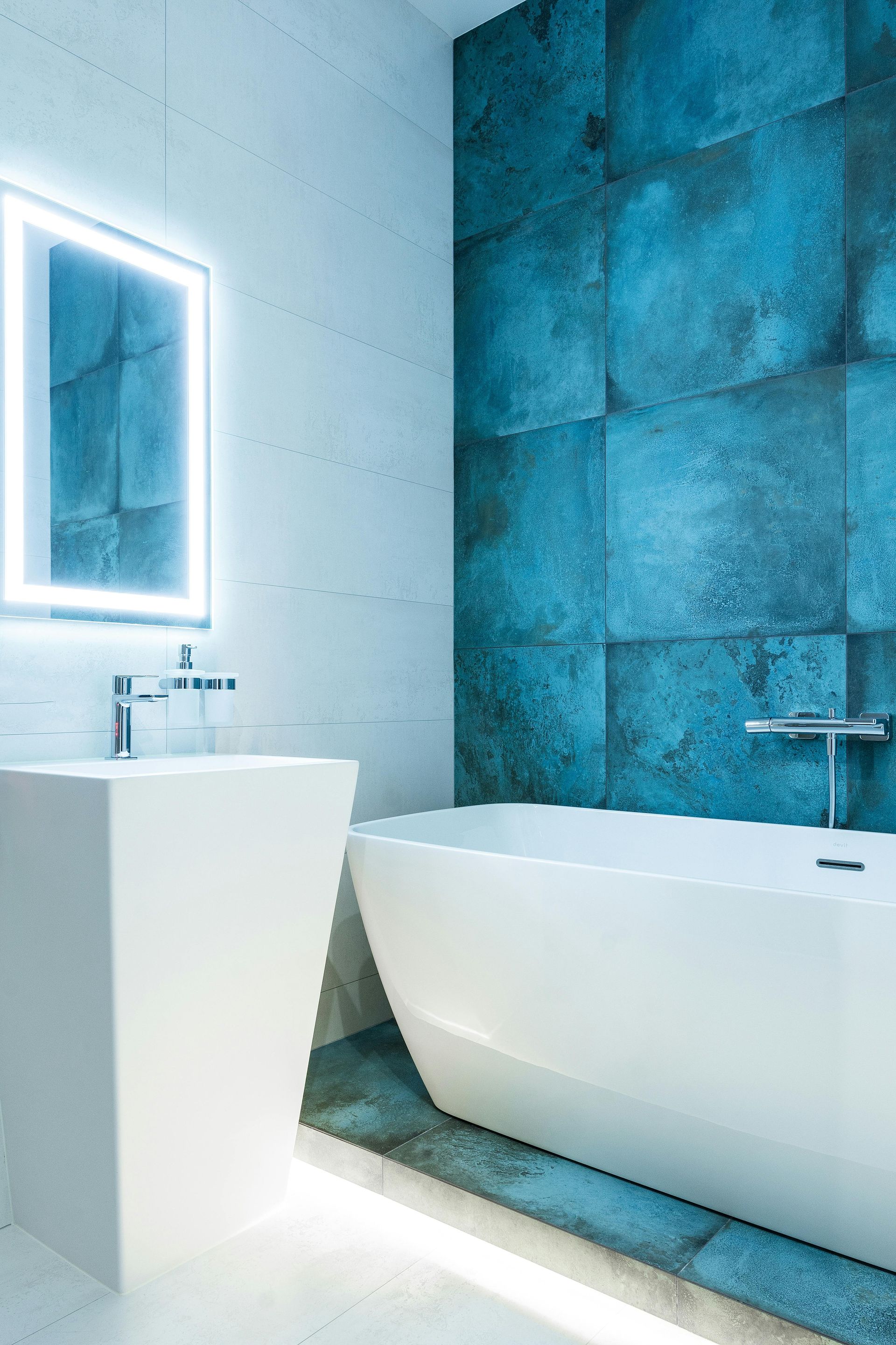 Modern bathroom with blue tile accent wall, white pedestal sink, and bathtub. Lit mirror.