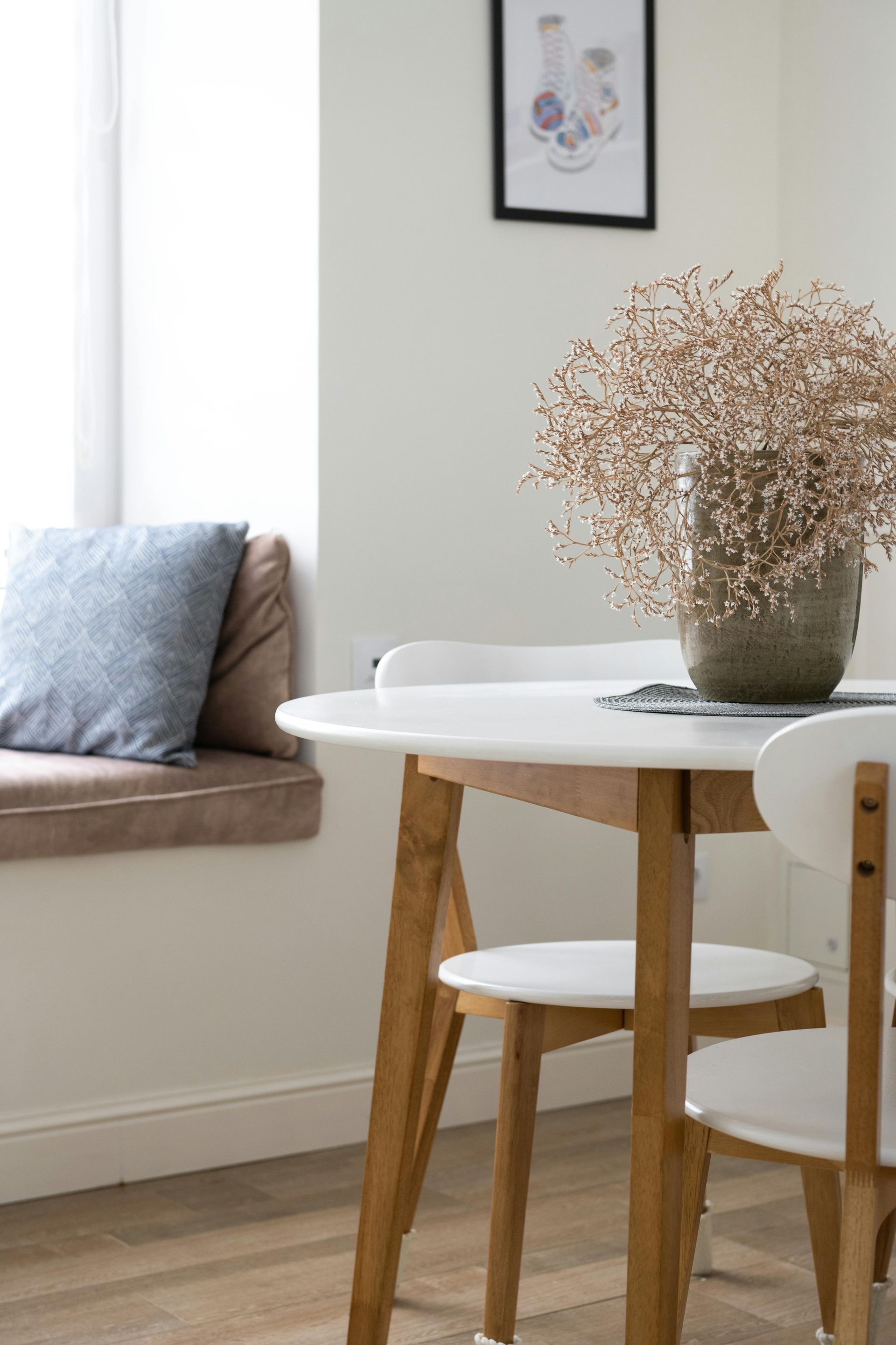 Round white table with wooden legs and white chairs, topped with vase of dried flowers, near a window seat.
