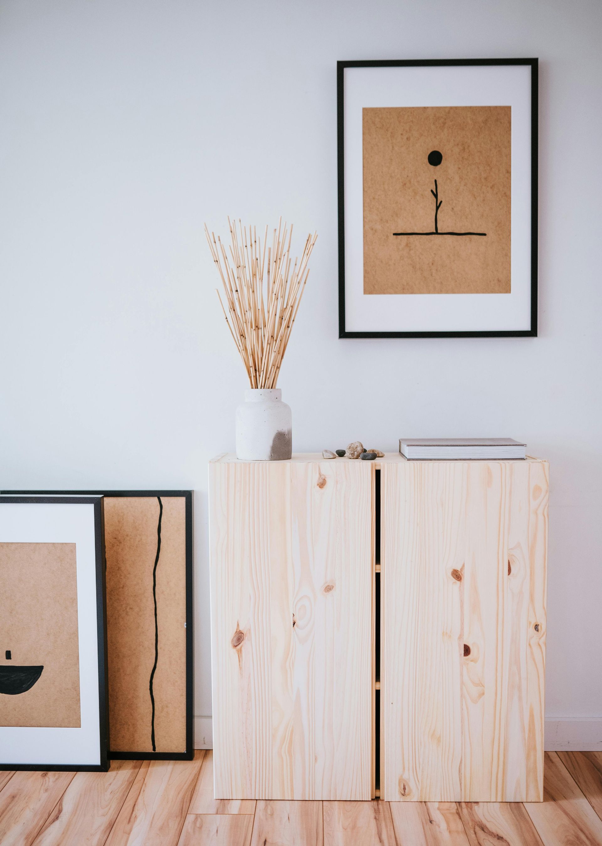 Wooden cabinet with art prints and vase of dried flowers against a white wall and wood floor.