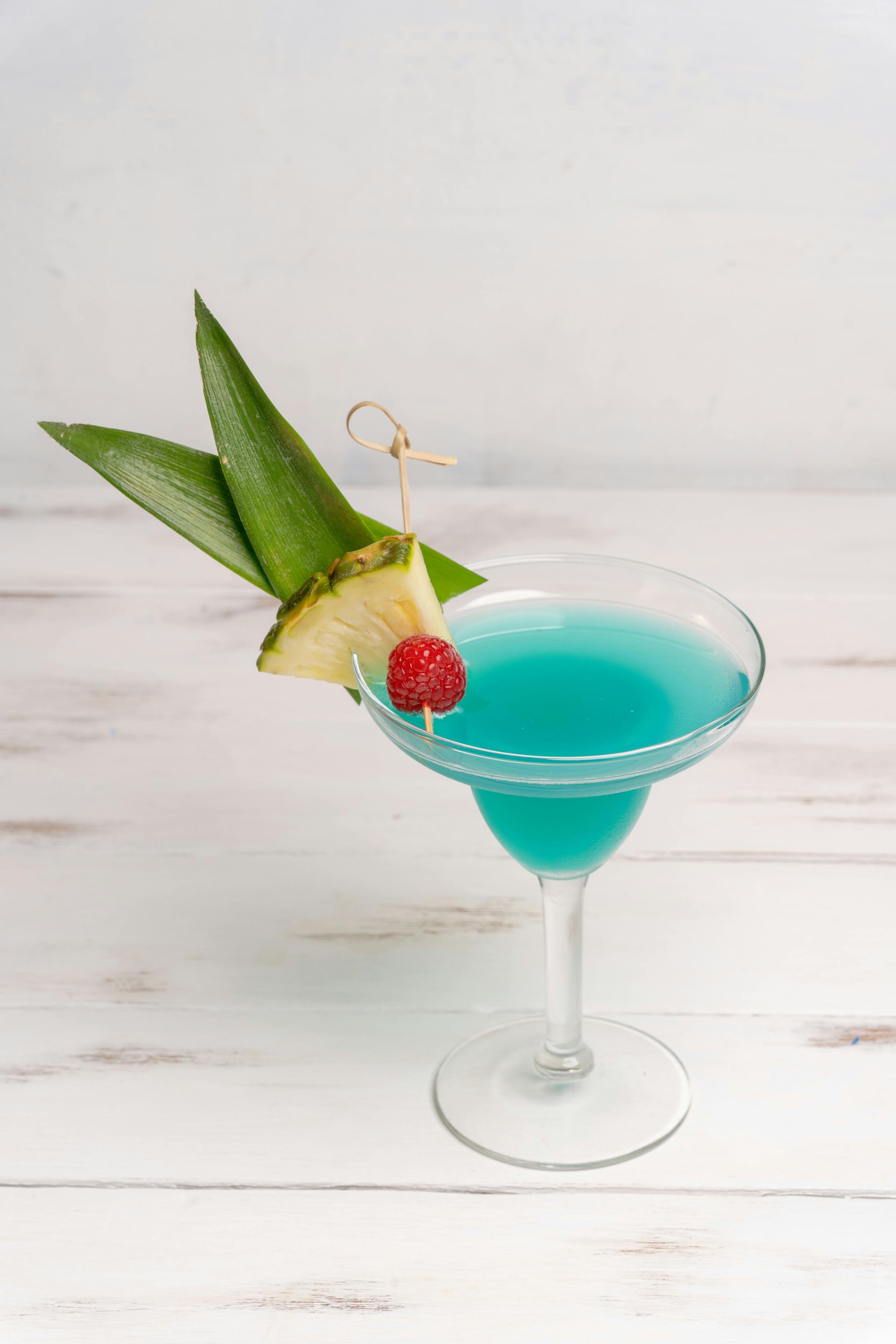 Blue cocktail in a margarita glass, garnished with pineapple, raspberry, and green leaves.