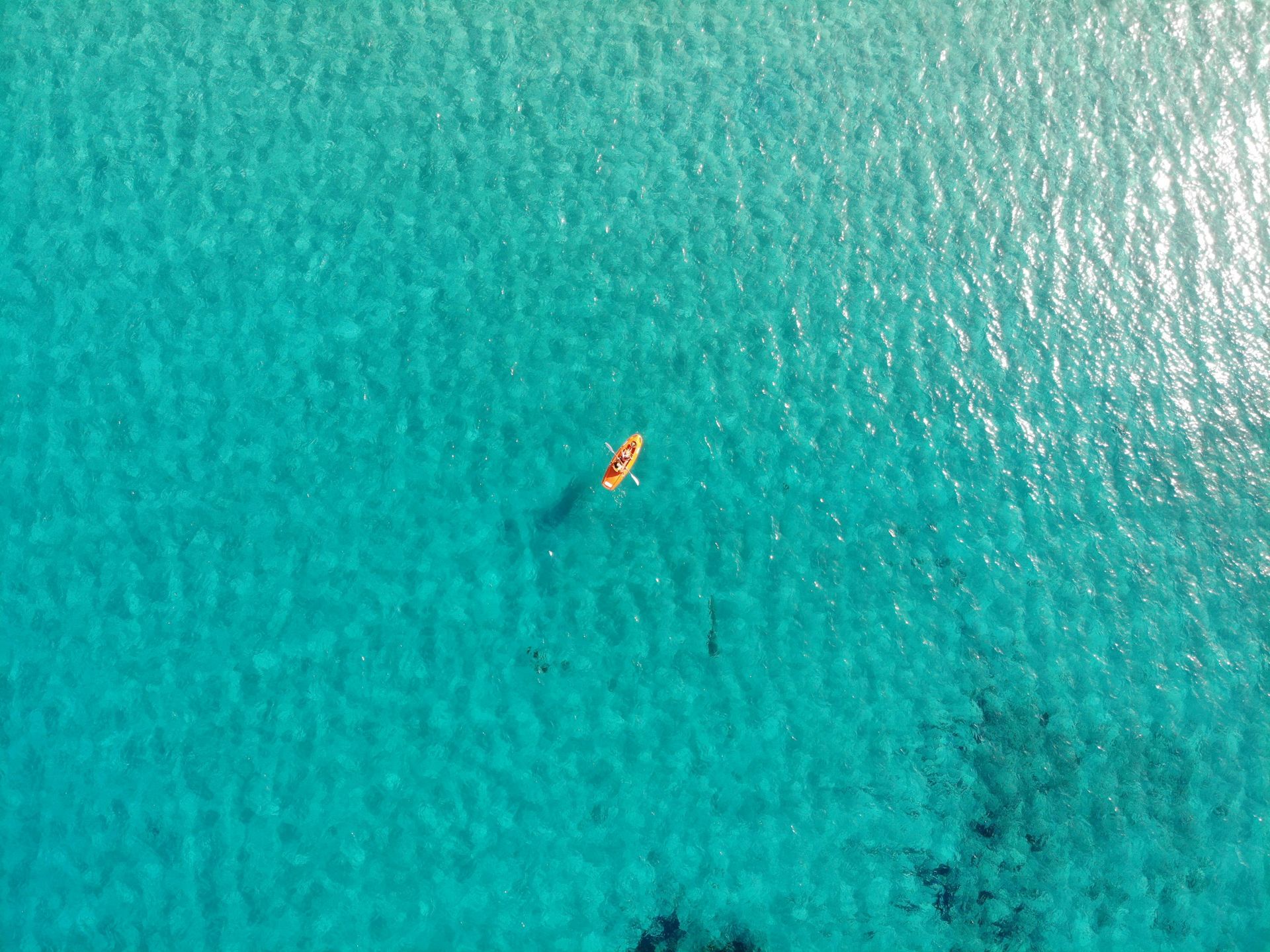 A small, light-colored kayak floats on clear, bright turquoise water, viewed from a high overhead drone perspective.