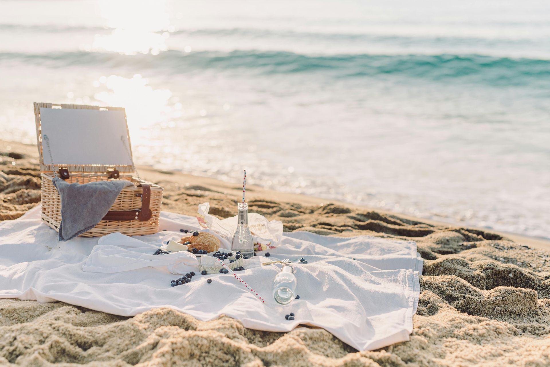 Picnic setup on a sandy beach. Wicker basket, champagne, and snacks on a white cloth near the ocean.