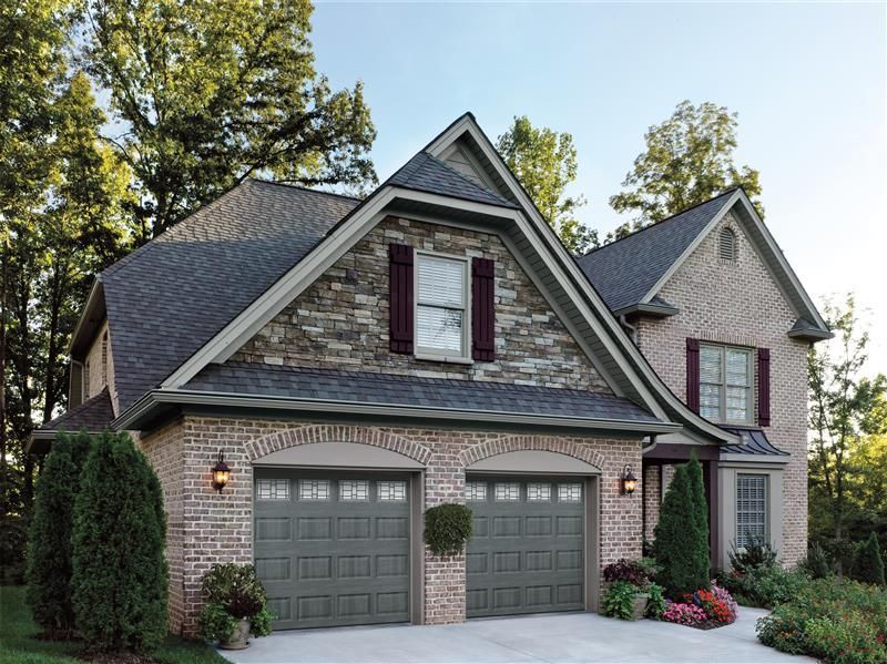 Two-story brick house with green garage doors, stone accents, and dark roof; surrounded by trees and landscaping.