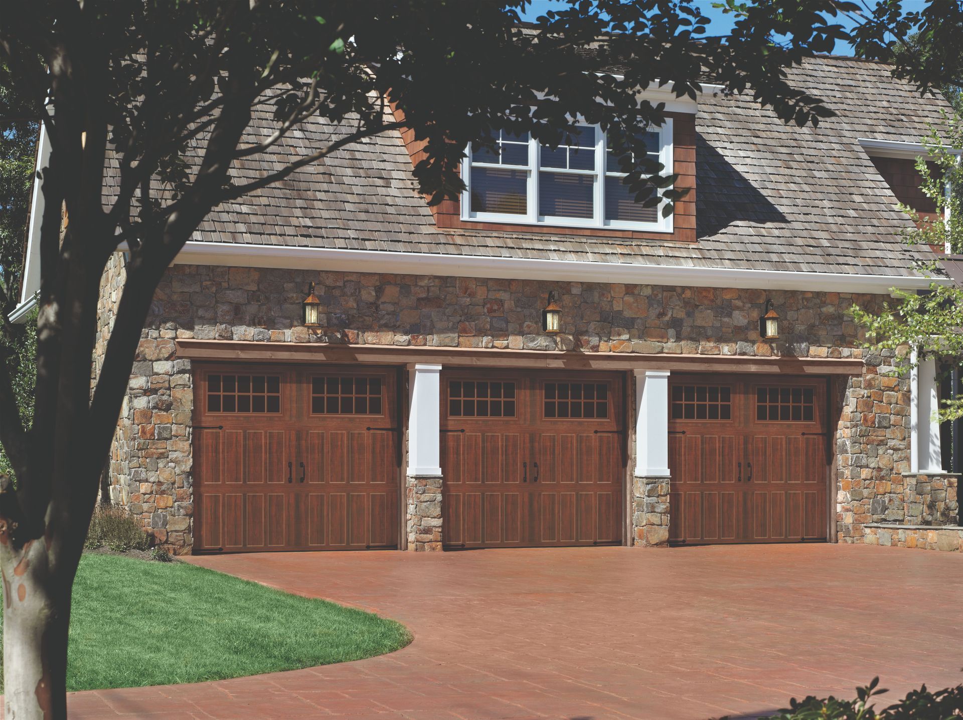 Three-car garage with brown wooden doors, stone facade, and a red brick driveway.