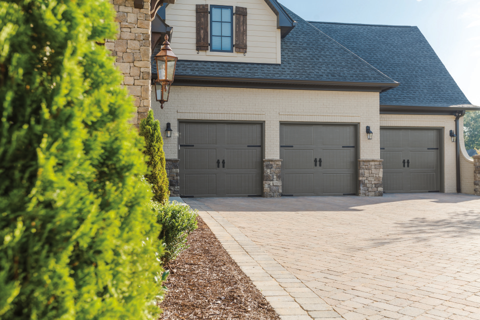 Beige three-car garage with dark brown accents and stone columns, on a brick paved driveway.