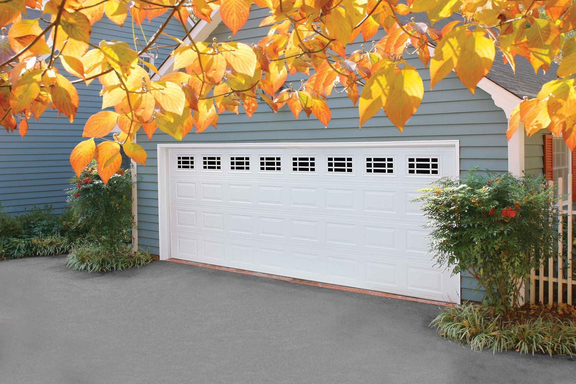 White garage door on a gray house, framed by orange autumn leaves and shrubs.