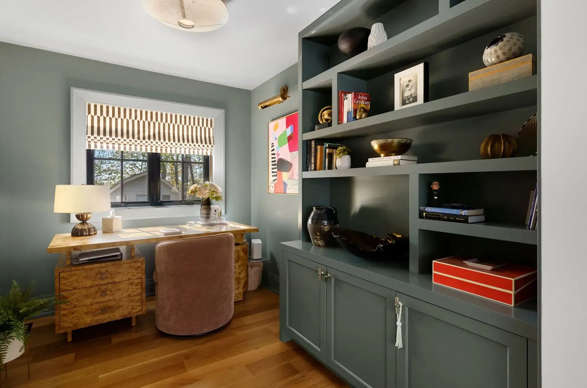 Home office with desk, lamp, window, woven chair, and built-in dark green shelves with books and decor.