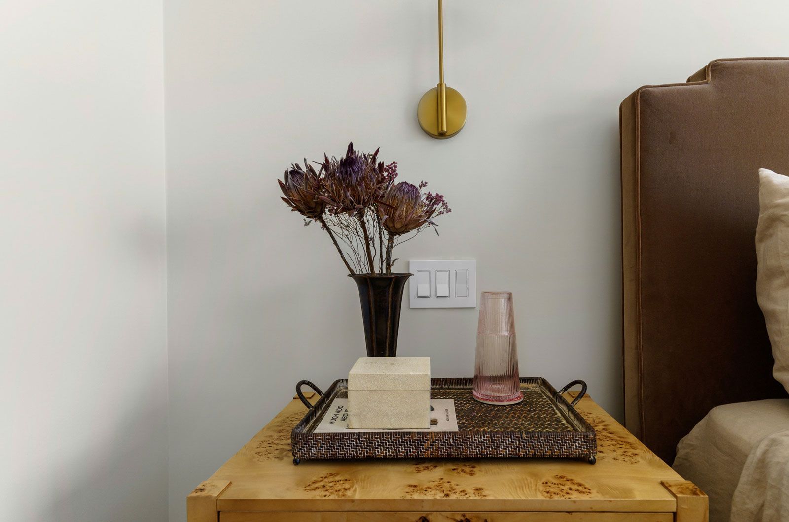 Bedside table with vase of dried flowers, tray, lamp, and candle beside a beige bed in a neutral room