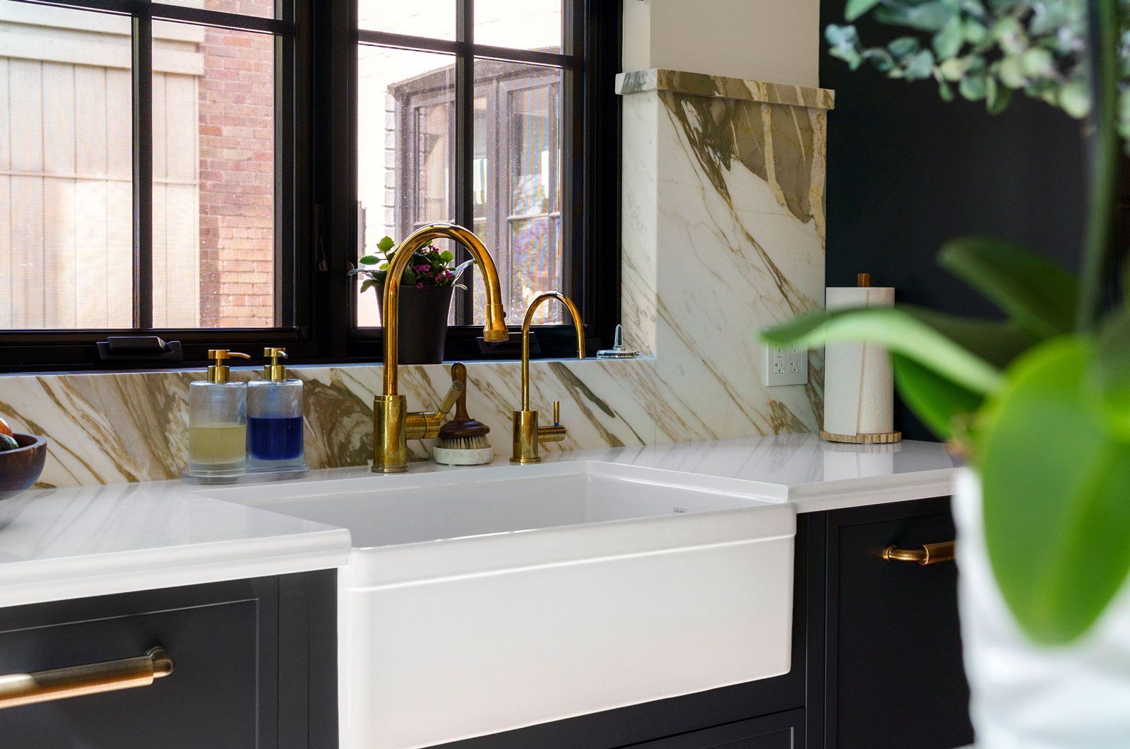 Modern kitchen sink with gold faucet, marble backsplash, black cabinets, and a blurred green plant foreground