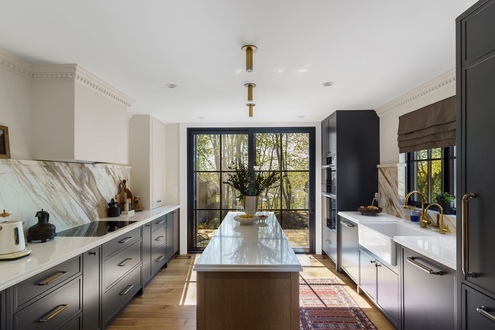 Modern kitchen with marble island, dark cabinetry, gold fixtures, and large glass doors to a garden.