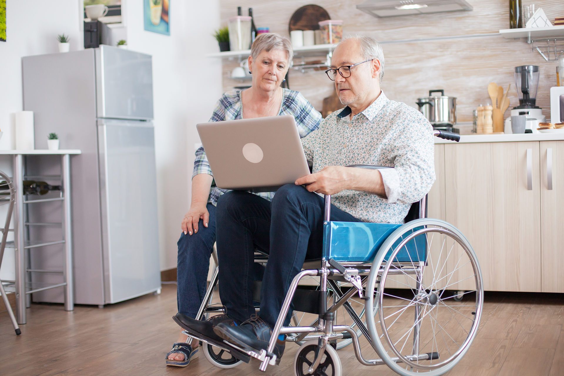 A man in a wheelchair is using a laptop computer while a woman sits next to him.