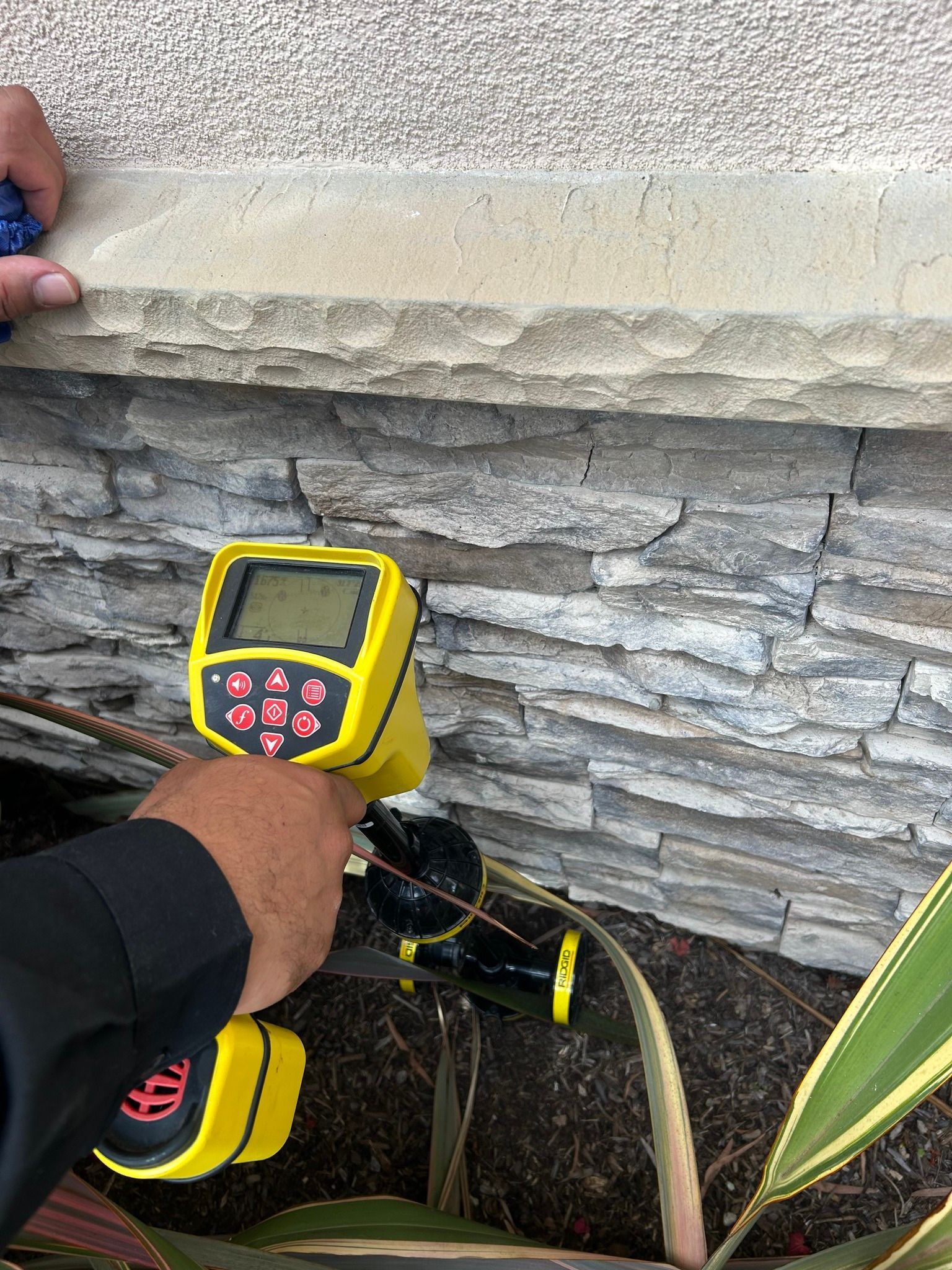 Person using a yellow thermal camera to inspect stone wall near landscaping plants