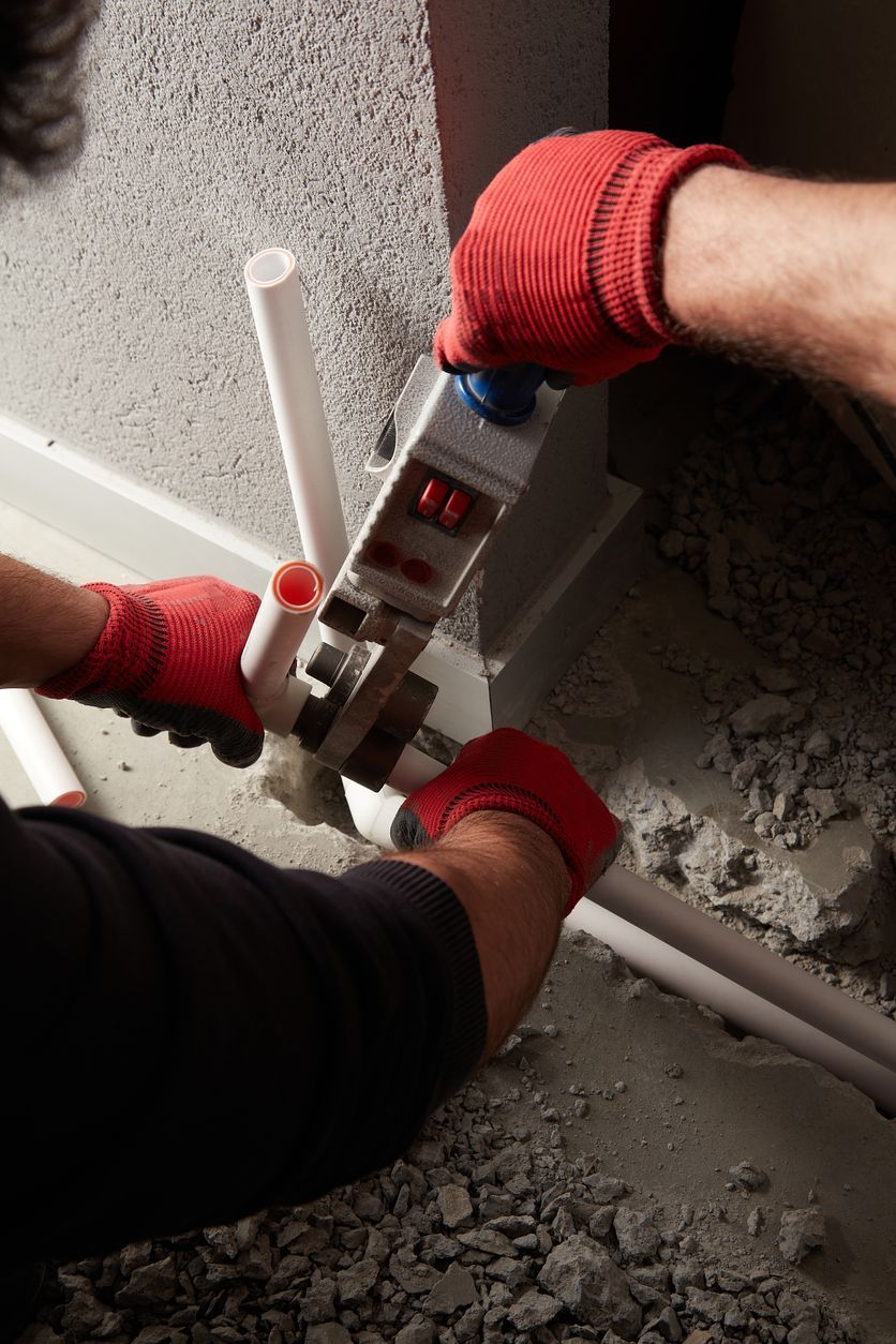 Gloved hands use a pipe cutter on a white PVC pipe amid gravel and concrete debris.