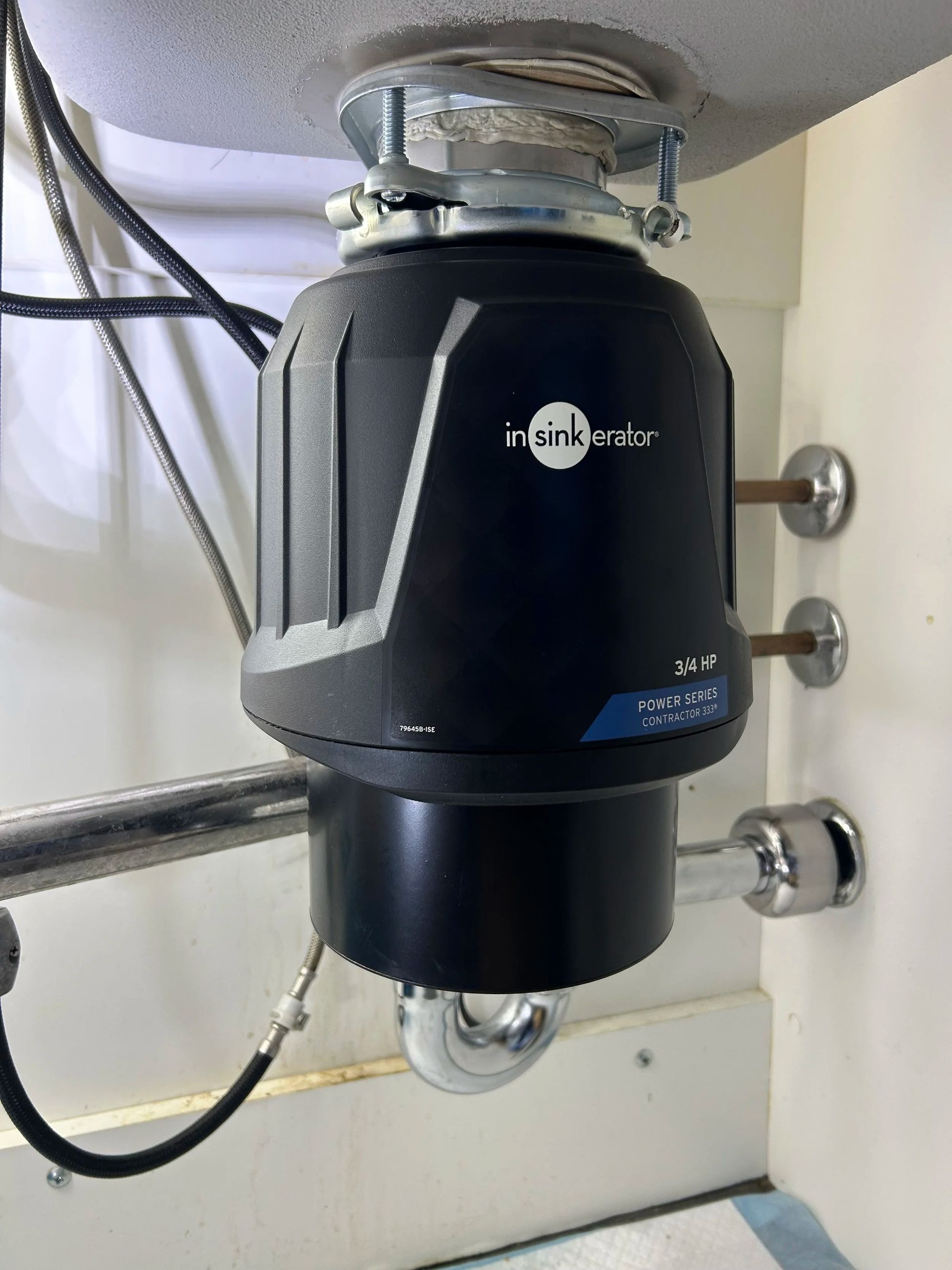 Black under-sink garbage disposal unit mounted beneath a sink with pipes and wiring visible