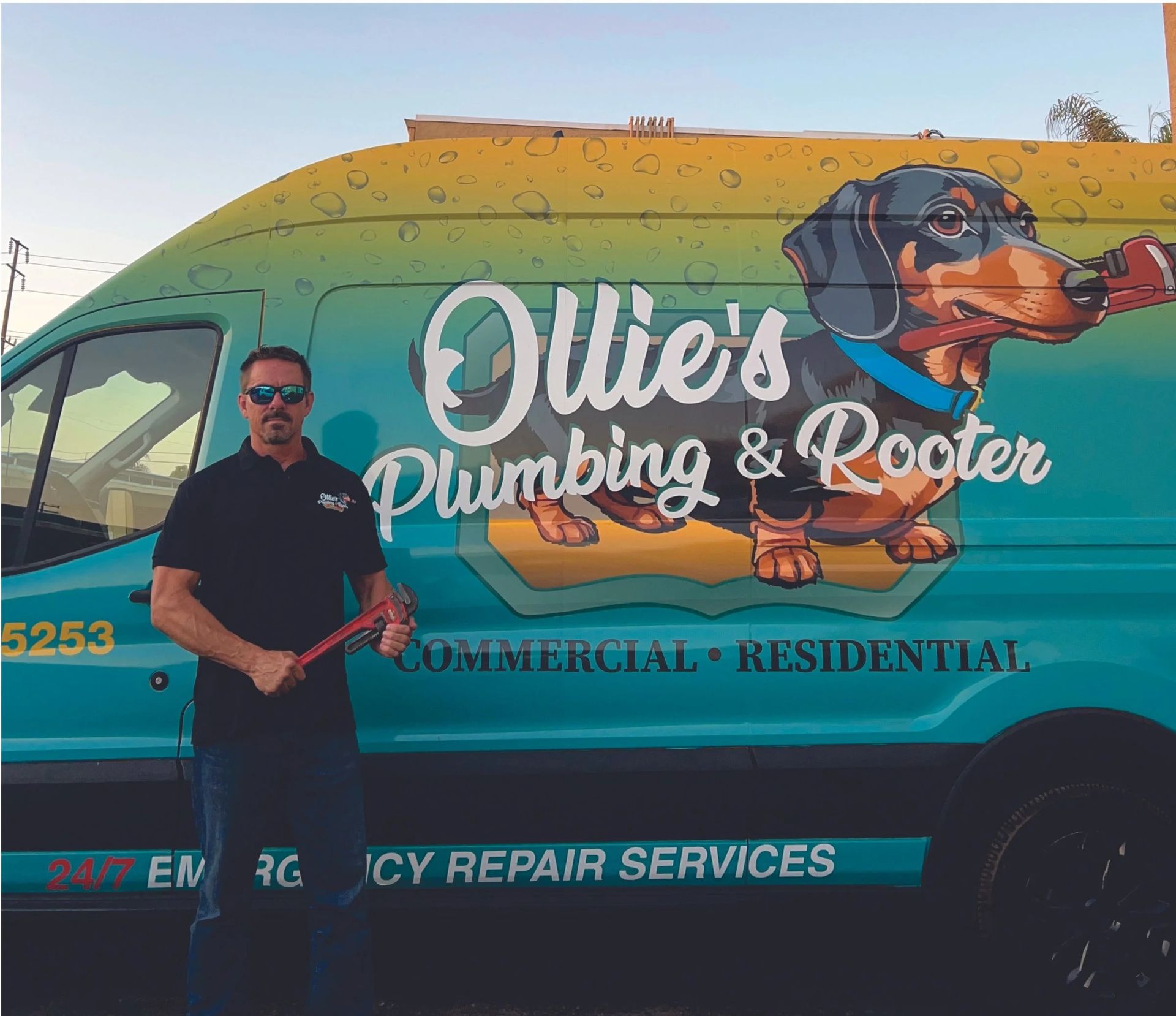 Man standing beside a colorful Ollie’s Plumbing & Rooter service van with dog logo and “Commercial Residential” text