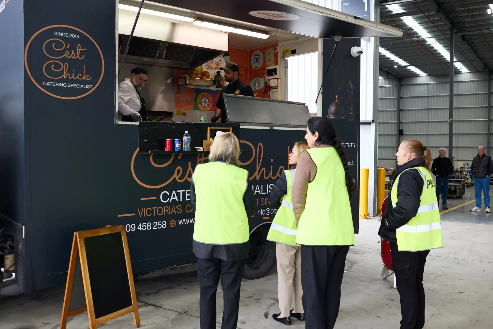 A group of people are gathered under a tent with a sign that says Cest Chick Food truck catering