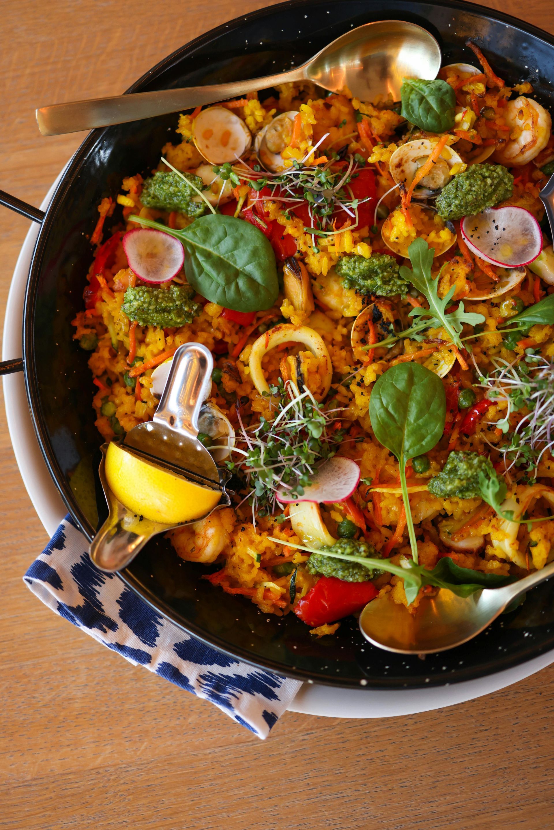 A pan of Paella and Spanish food with a spoon and a lemon on a table.