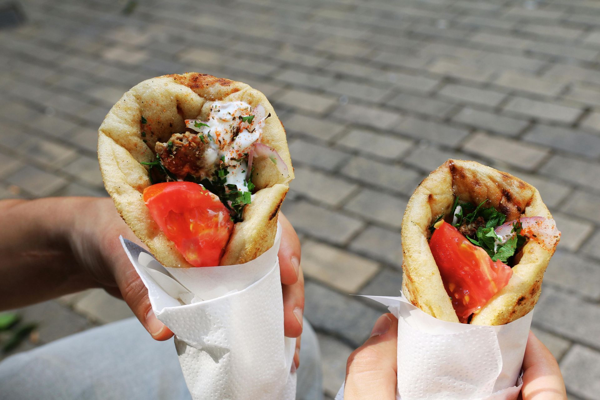 A person is holding two souvlaki in their hands on a brick footpath.