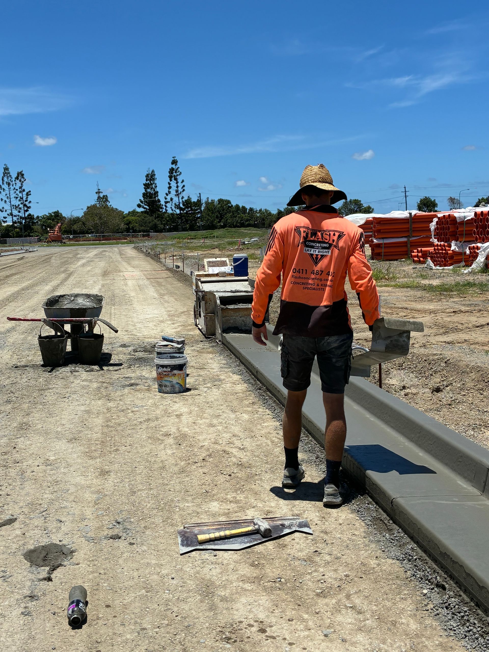 Construction worker in orange shirt walking beside a concrete curb at a roadwork site under a blue sky — Flash Concreting Pty Ltd In Nindaroo, QLD