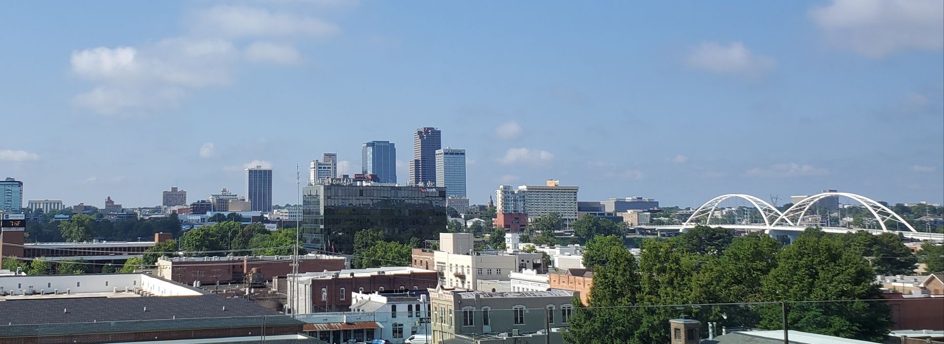 City skyline with tall buildings, a bridge, and trees under a blue sky.
