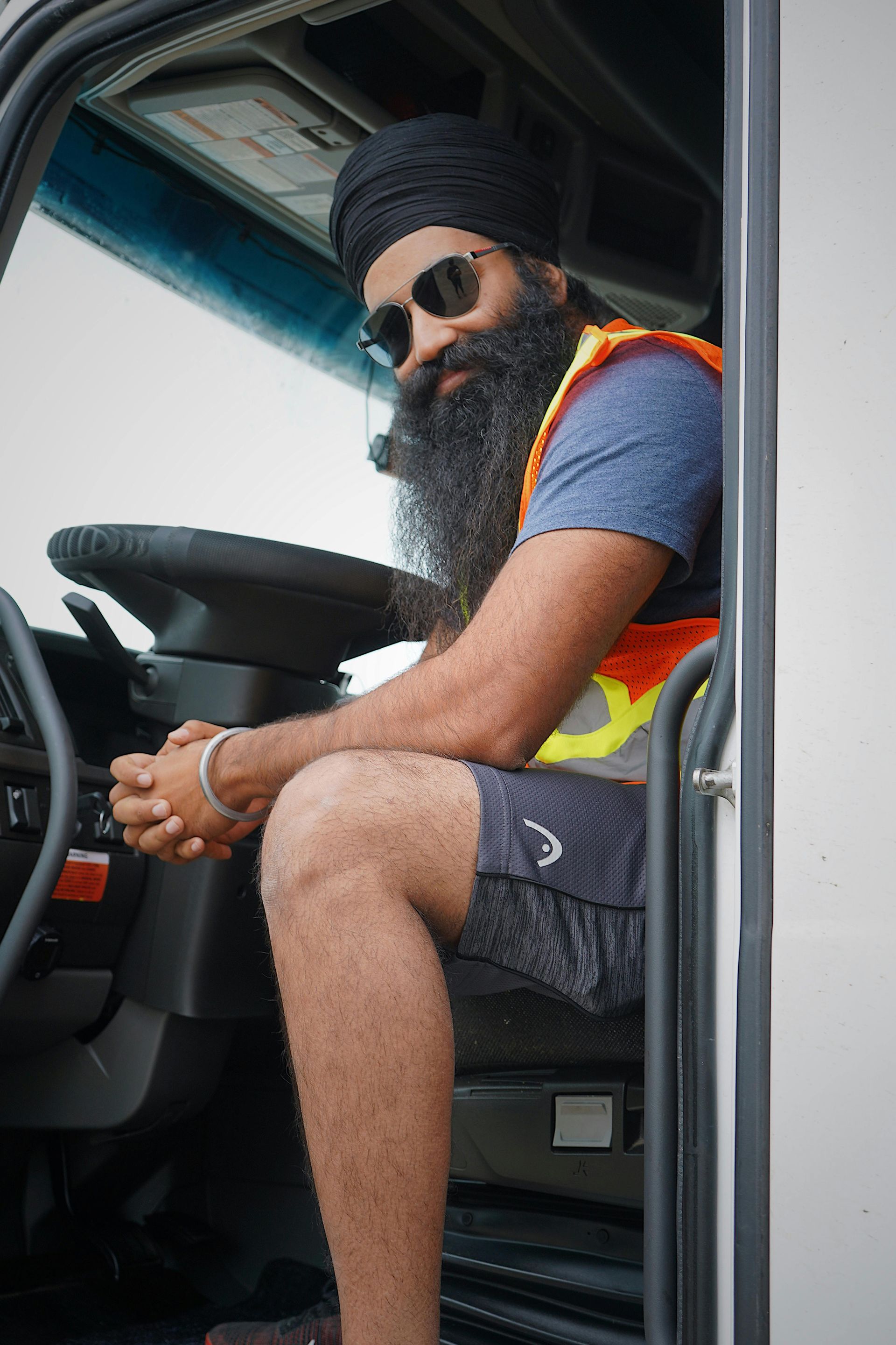 Man in turban and sunglasses sitting in a truck cabin, wearing a safety vest and shorts.