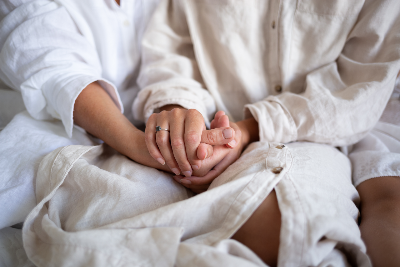 Two people in white clothing holding hands while seated on a bed