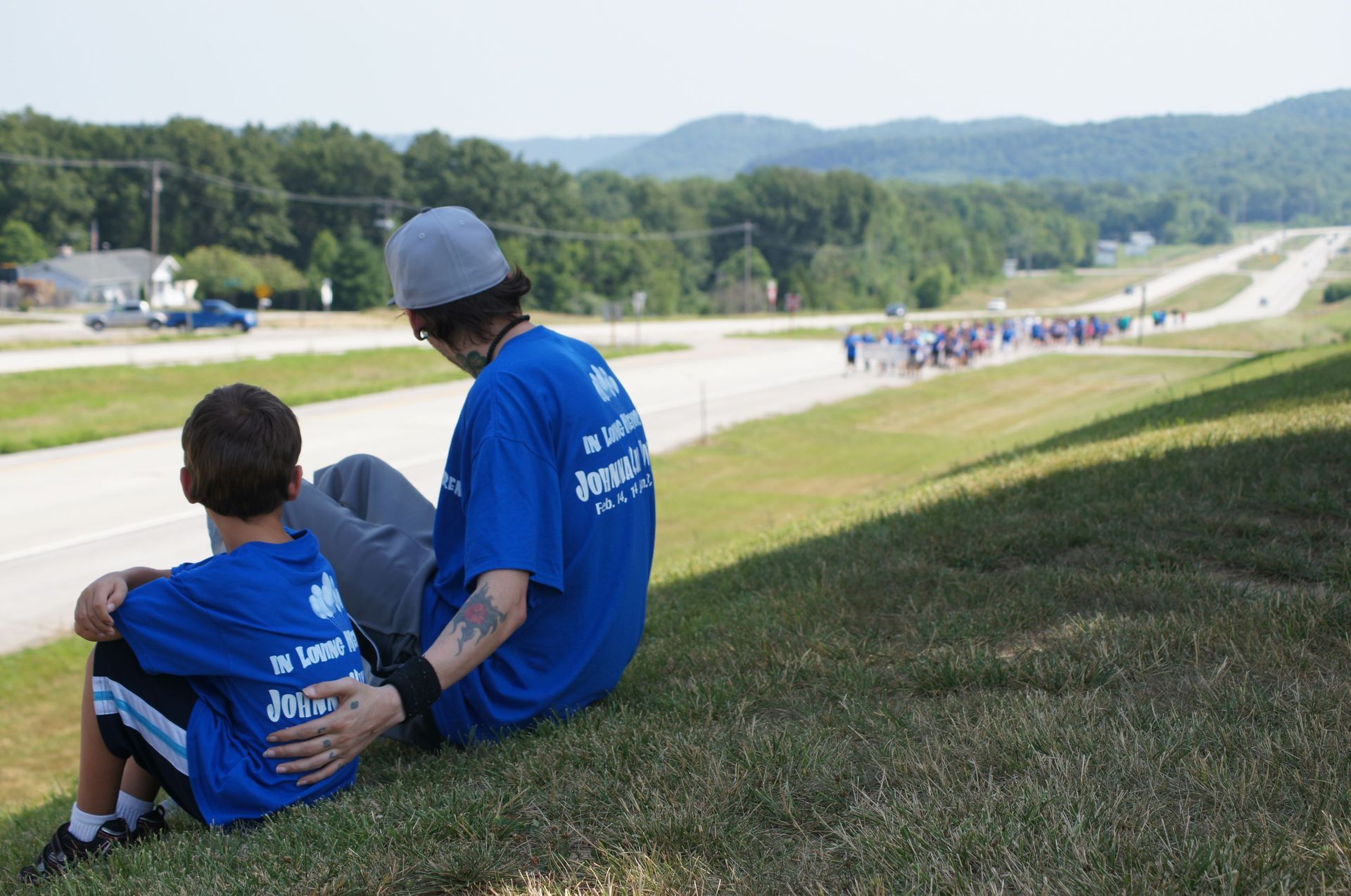 Two people in blue shirts sit on a grassy hill overlooking a road race route.