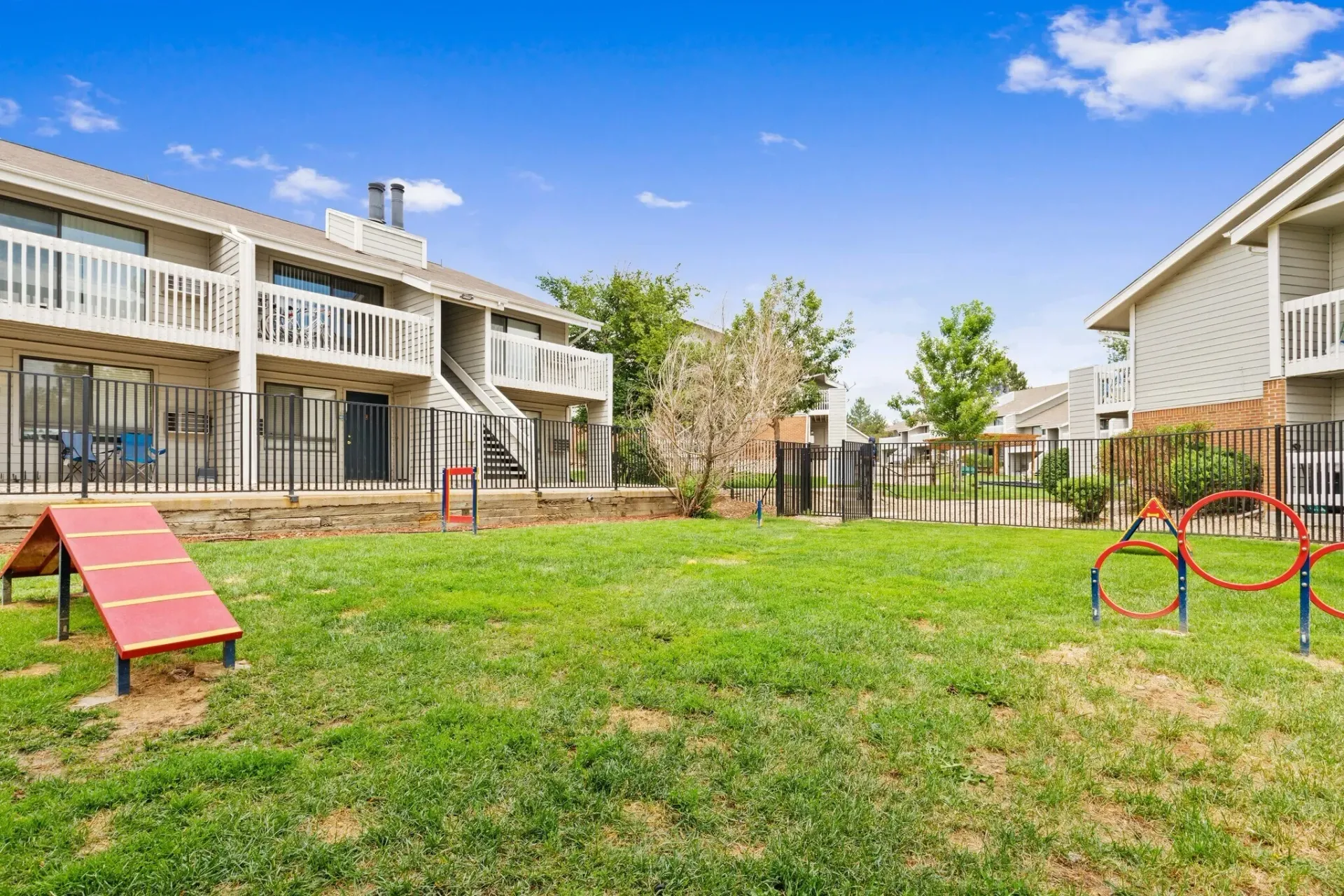 A fenced dog park with agility equipment like ramps and hoops, next to apartment buildings.