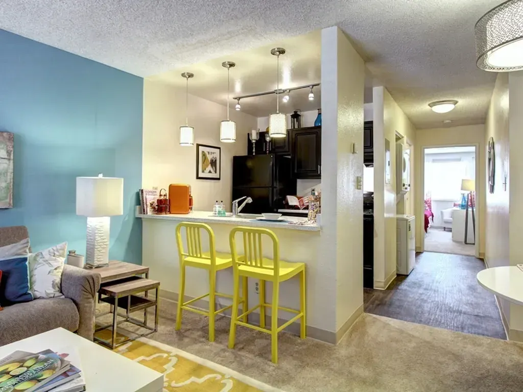 Open living room with kitchen bar, yellow stools, sofa, and hallway to a bedroom