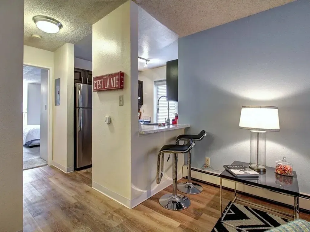 Modern apartment living area with a desk, lamp, stools, and kitchen in the background