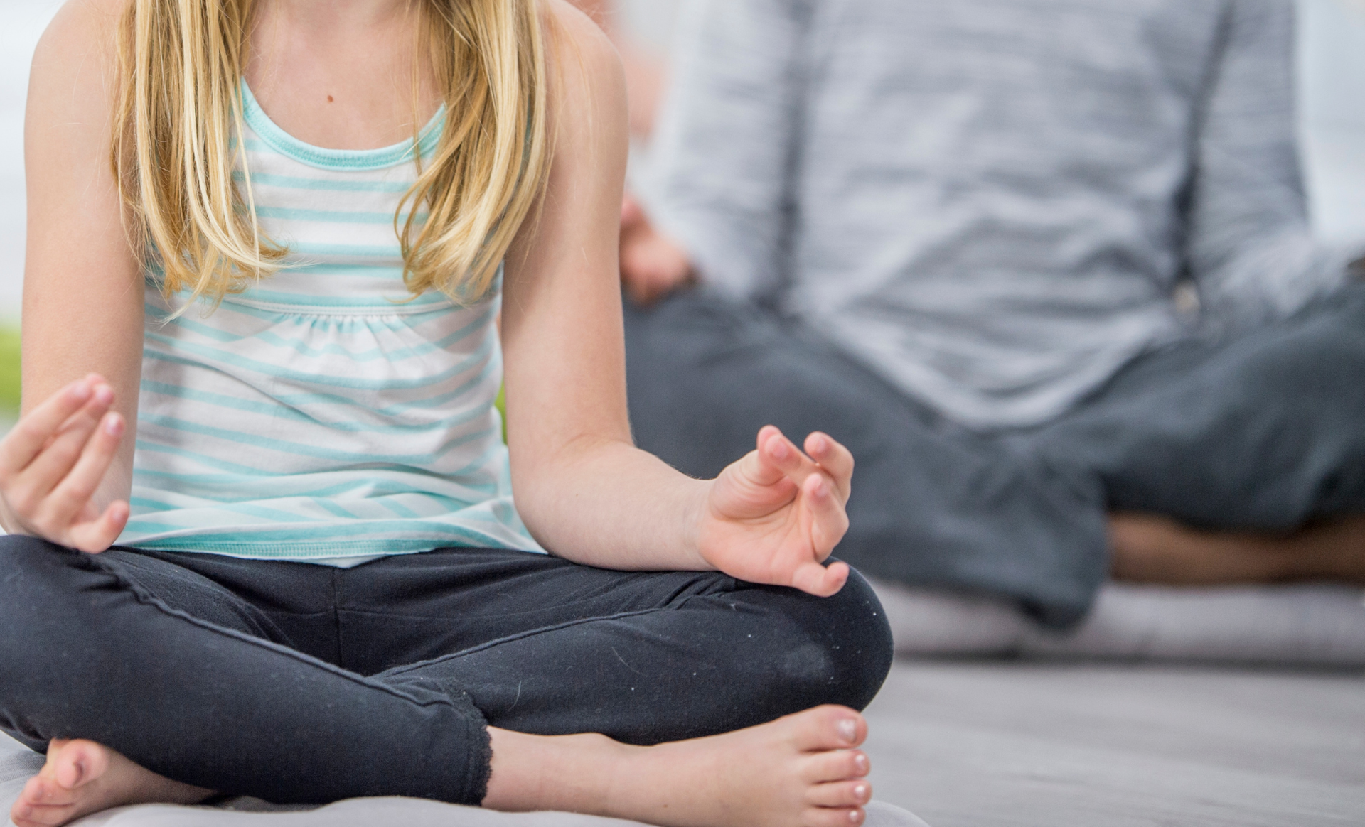 Child meditating cross-legged indoors, with another person blurred in the background