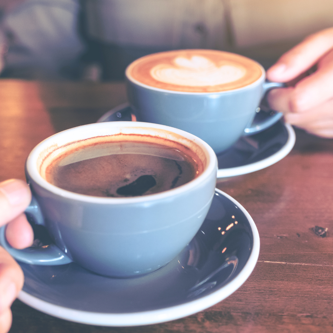 Two blue coffee cups on saucers on a wood table, one black coffee, one latte. Hands hold the cups.
