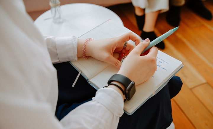 Person writes in notebook, holding pen with both hands, wearing watch and bracelet, close-up shot.