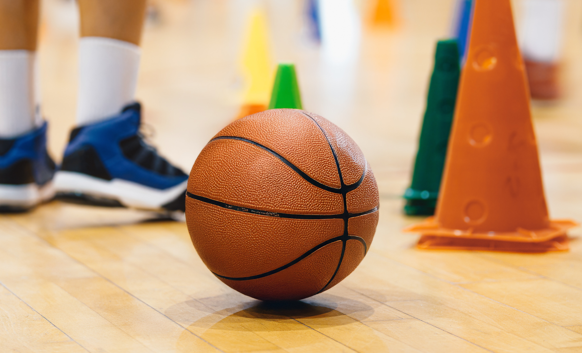 Basketball on a gym floor with blurred cones and players’ shoes in the background