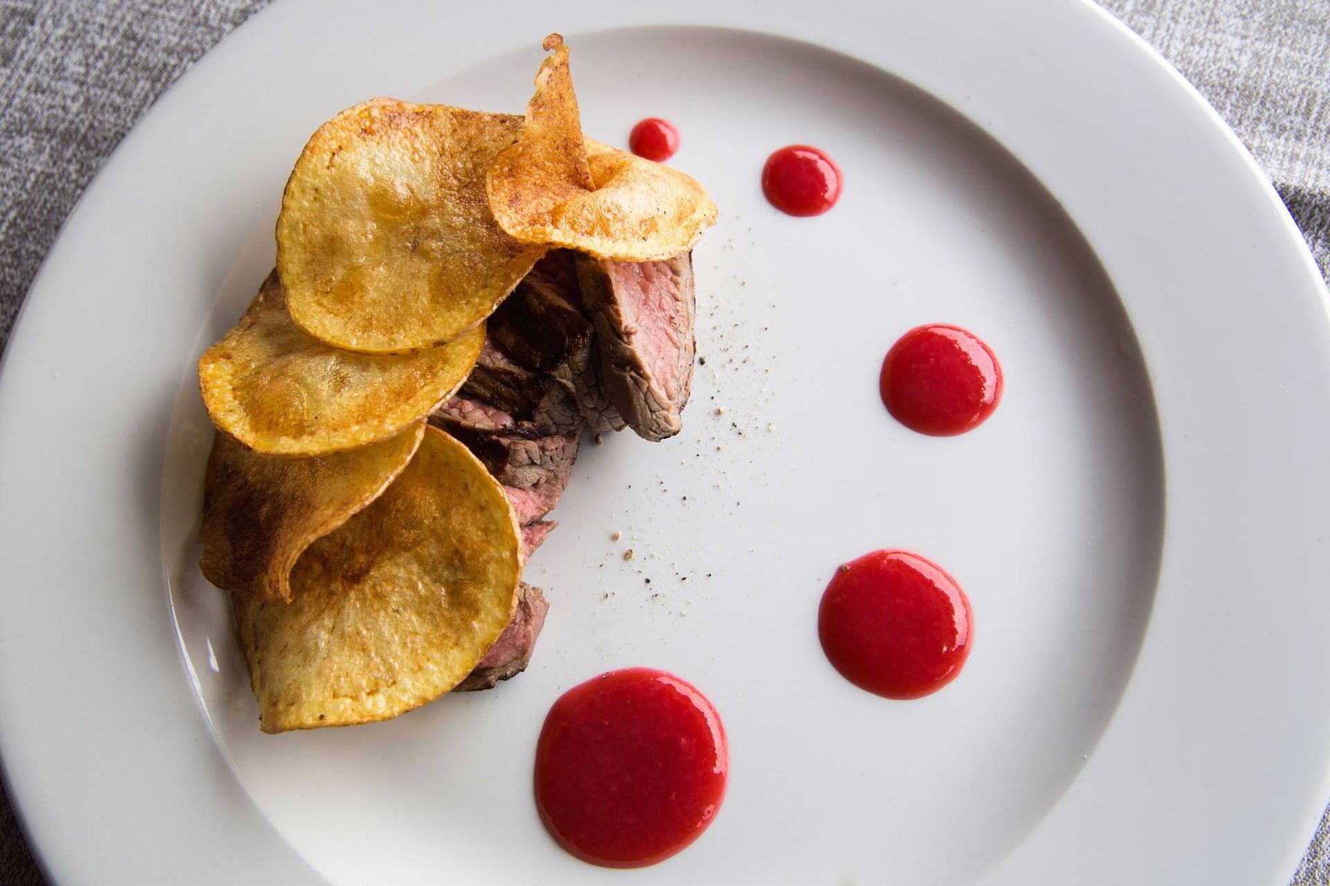 Piatto gourmet impiattato con carne affettata, patatine croccanti e gocce di salsa rossa su un piatto bianco.