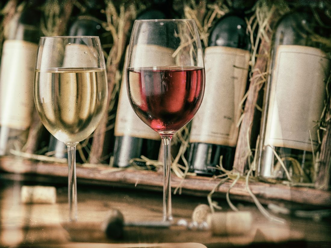 Due calici da vino, uno bianco e uno rosso, davanti a bottiglie di vino su un tavolo di legno.