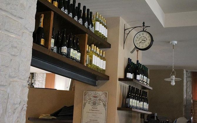 Bottiglie di vino esposte su scaffali a muro in un piccolo negozio con orologio a sospensione e ventilatore a soffitto.
