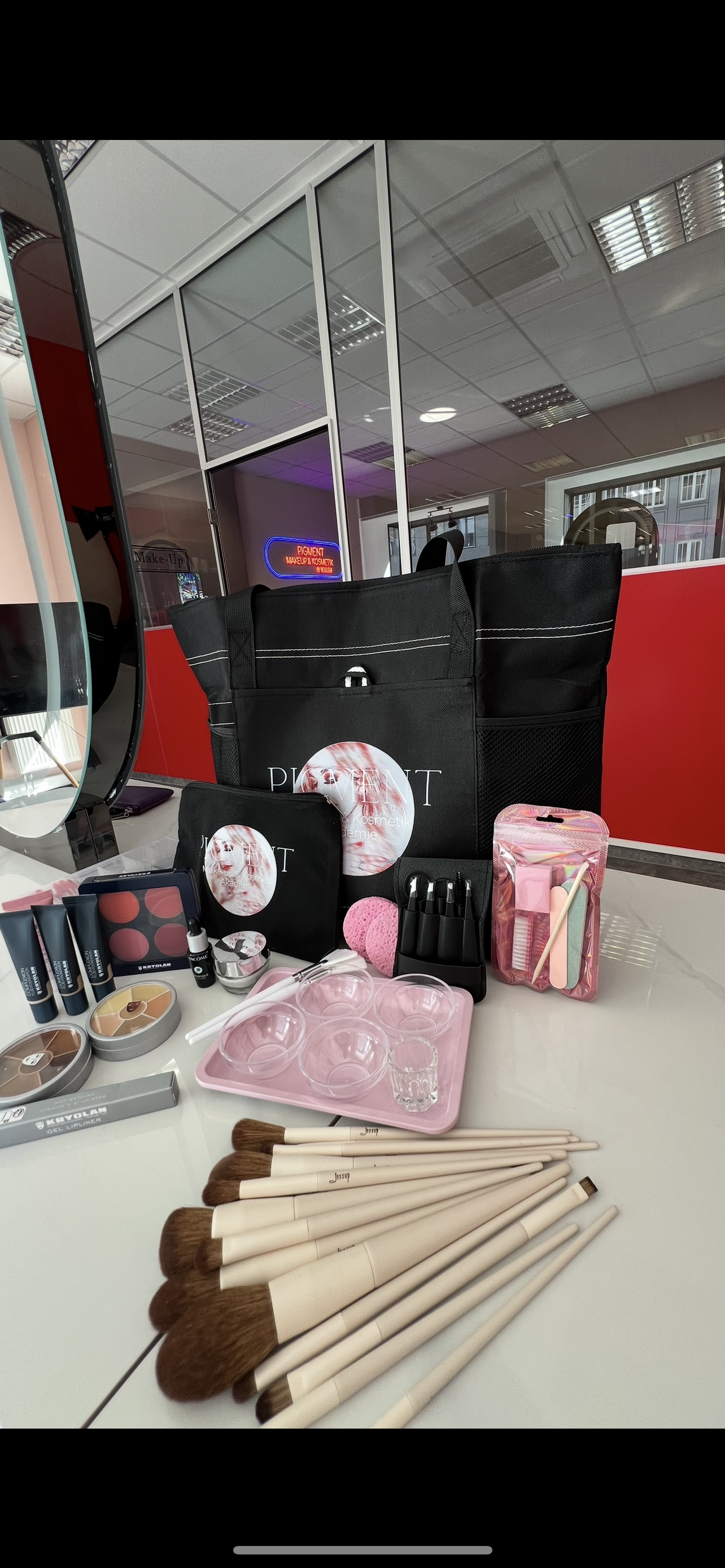 Auf einer Fläche sind Schminkpinsel und Kosmetikprodukte angeordnet. Im Hintergrund sind schwarze Taschen und Behälter zu sehen.