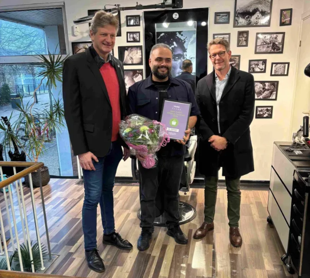 Tre personer poserer indendørs, den ene holder blomster og en plakette, foran en væg med indrammede fotos