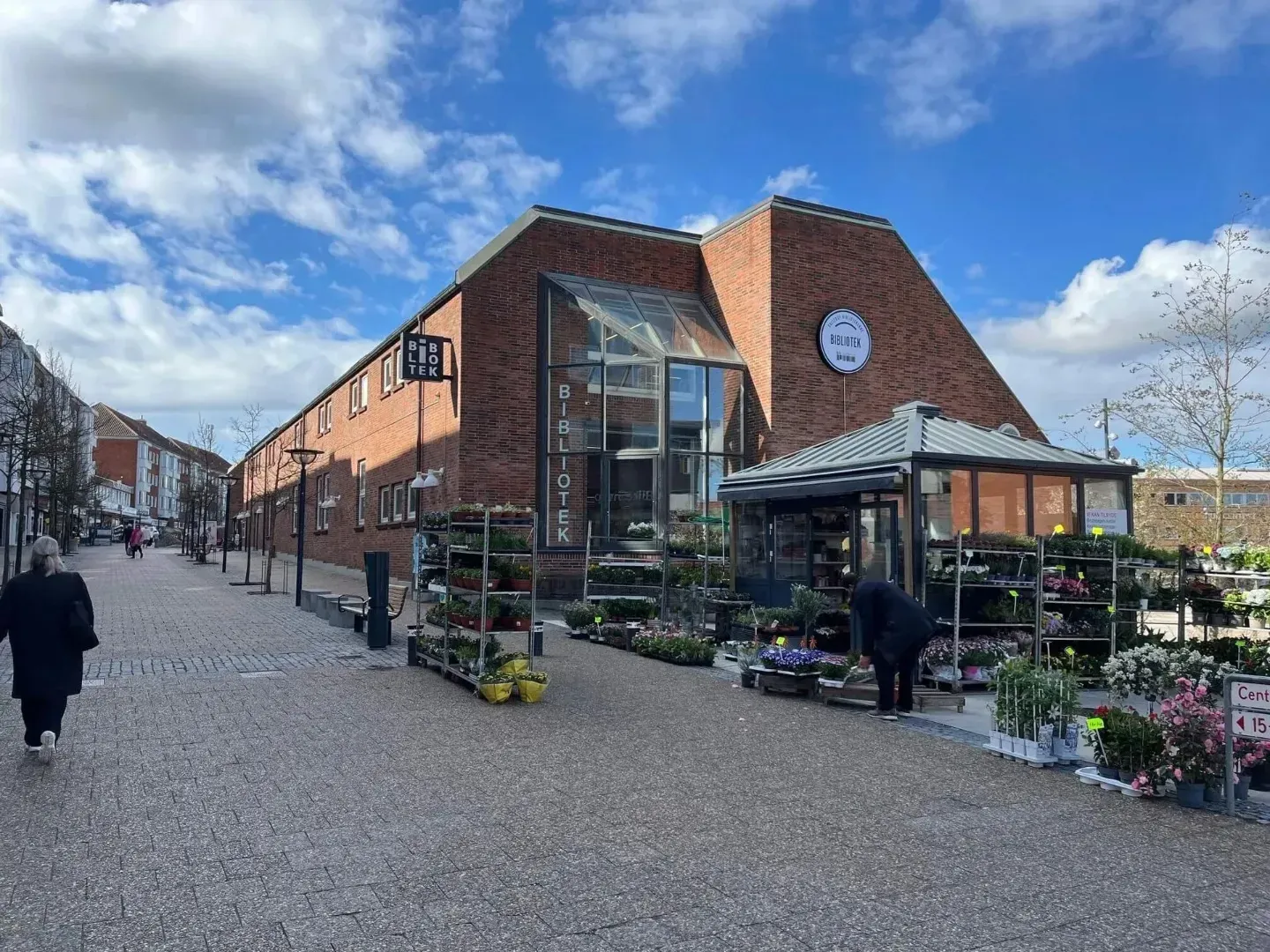 Murstensbutiksfacade med glasindgang og blomsterboder på en brostensbelagt gade under en overskyet blå himmel