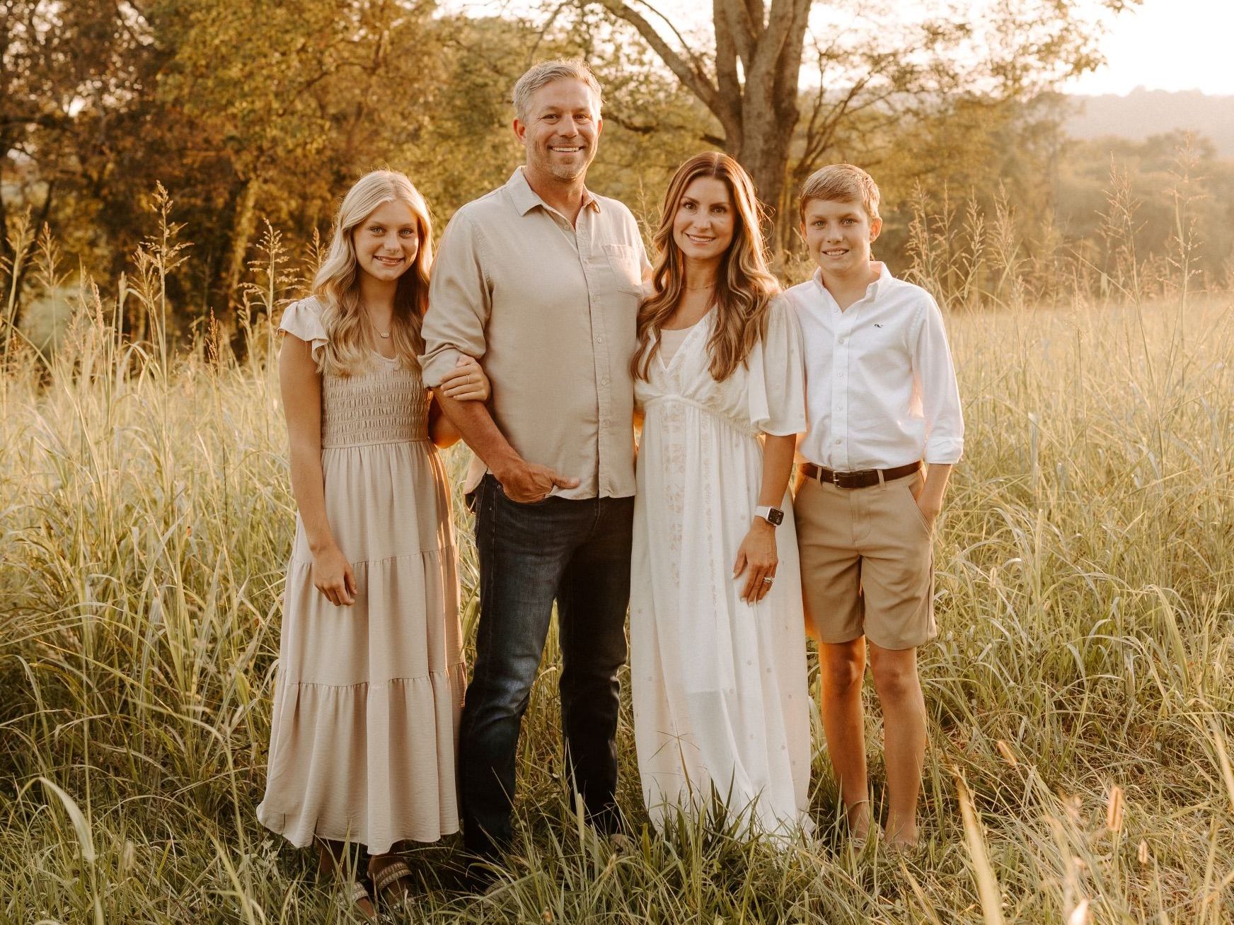 Family of four standing in a grassy field; sunlight, warm tones, two adults, two children.