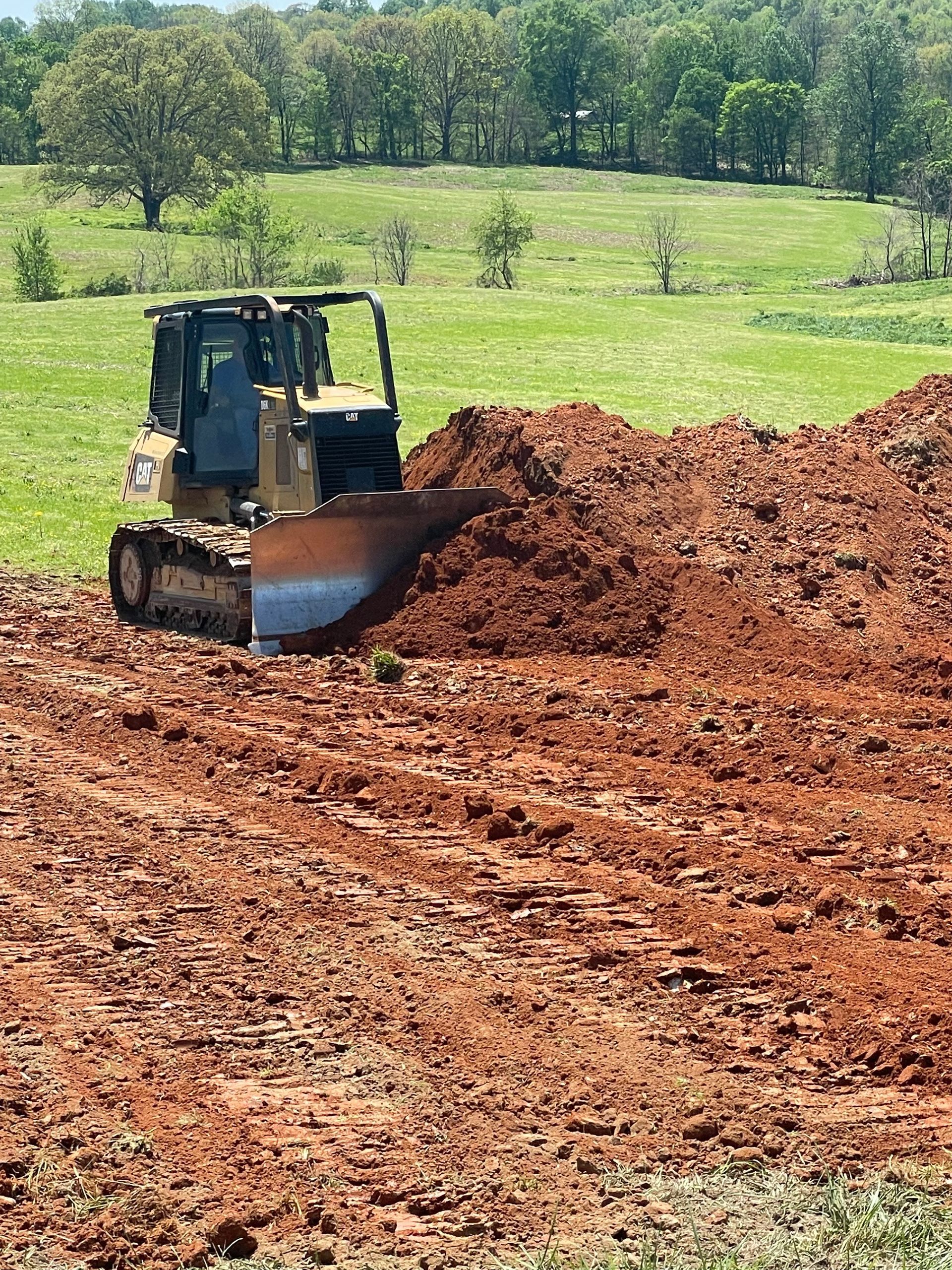 Bulldozer pushing a pile of red dirt on a muddy field, with trees and a green hill in the background.