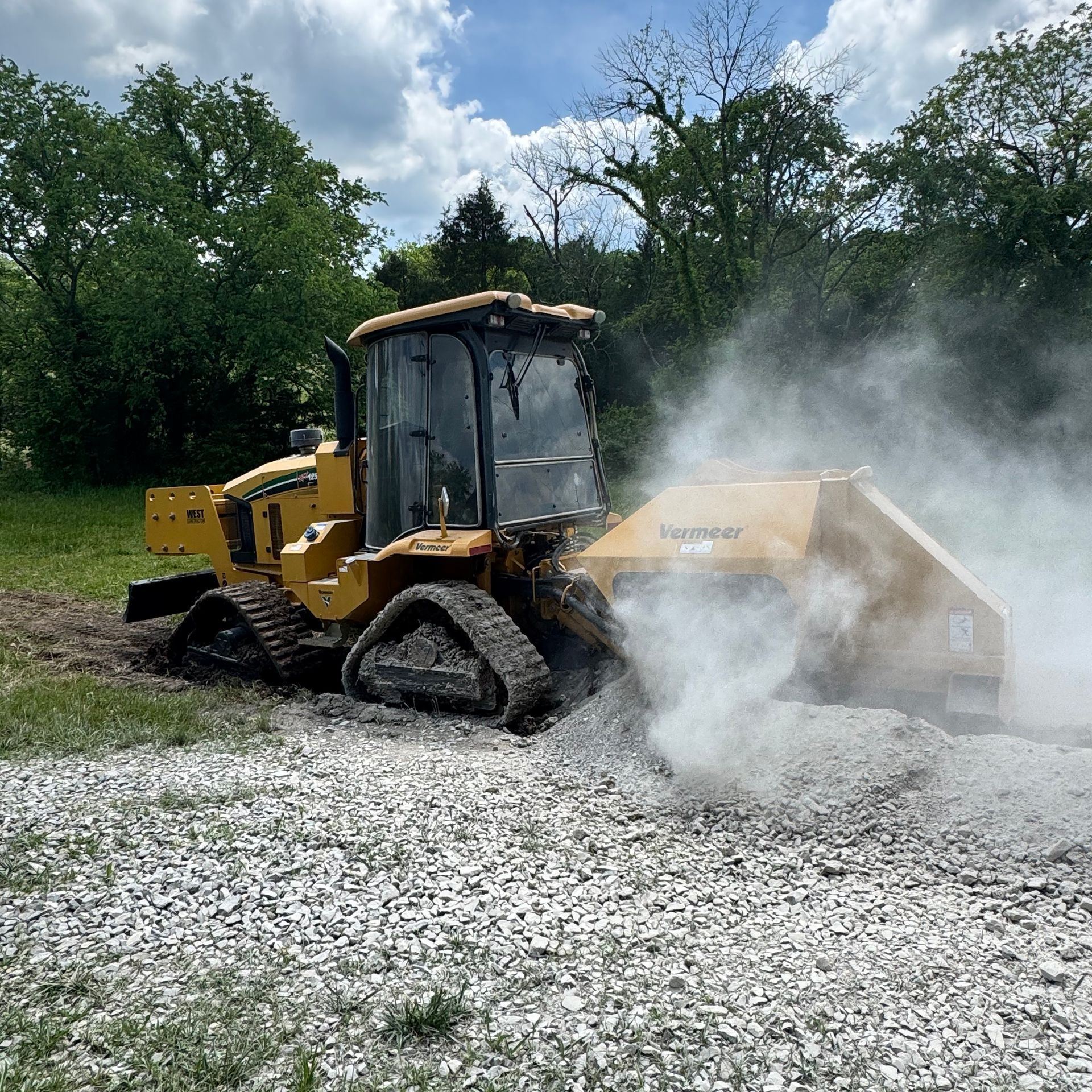 Yellow tracked machine grinding gravel, creating a cloud of dust outdoors.
