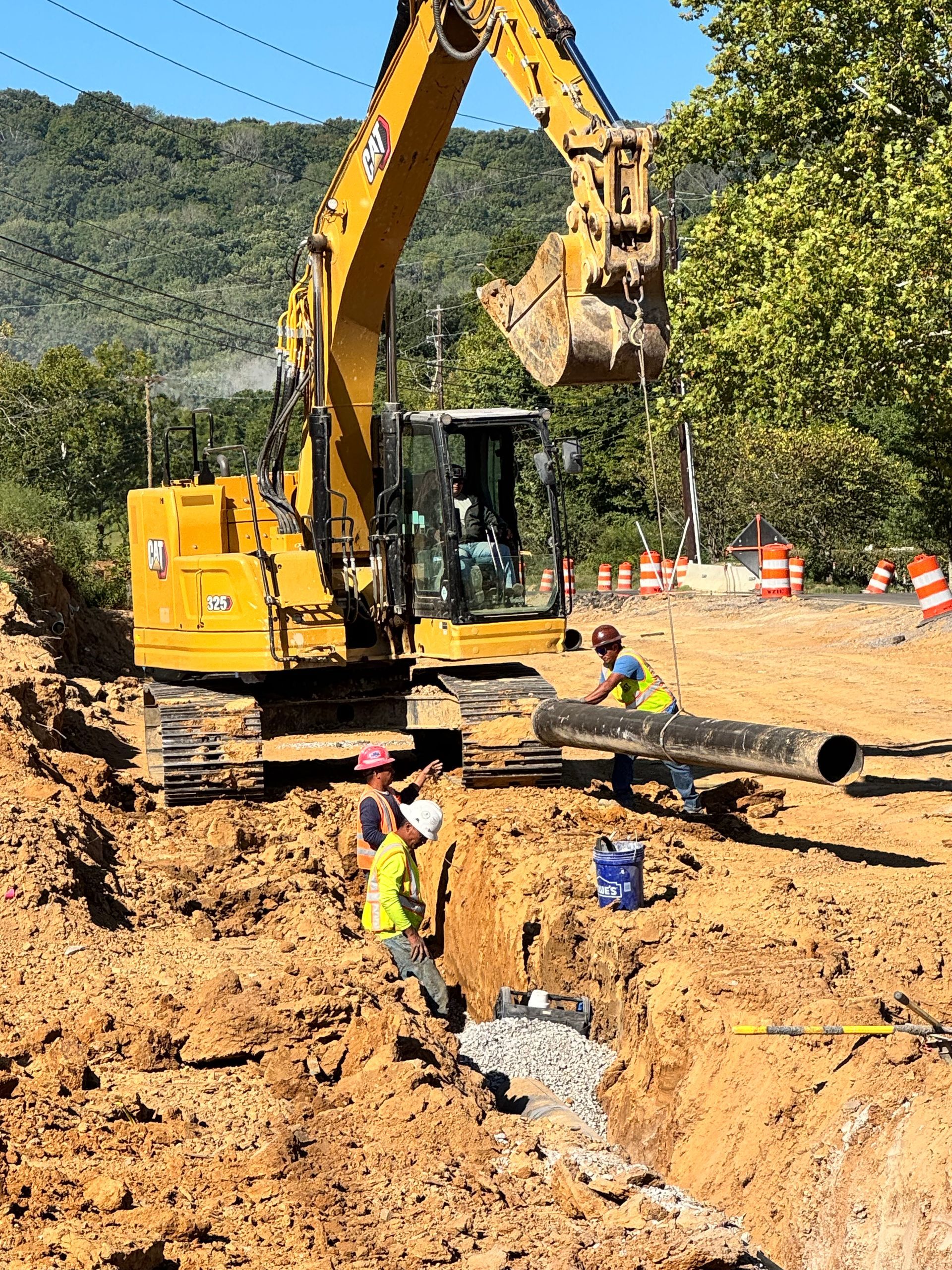 Construction workers in trench with excavator; setting pipe. Bright sunny day.