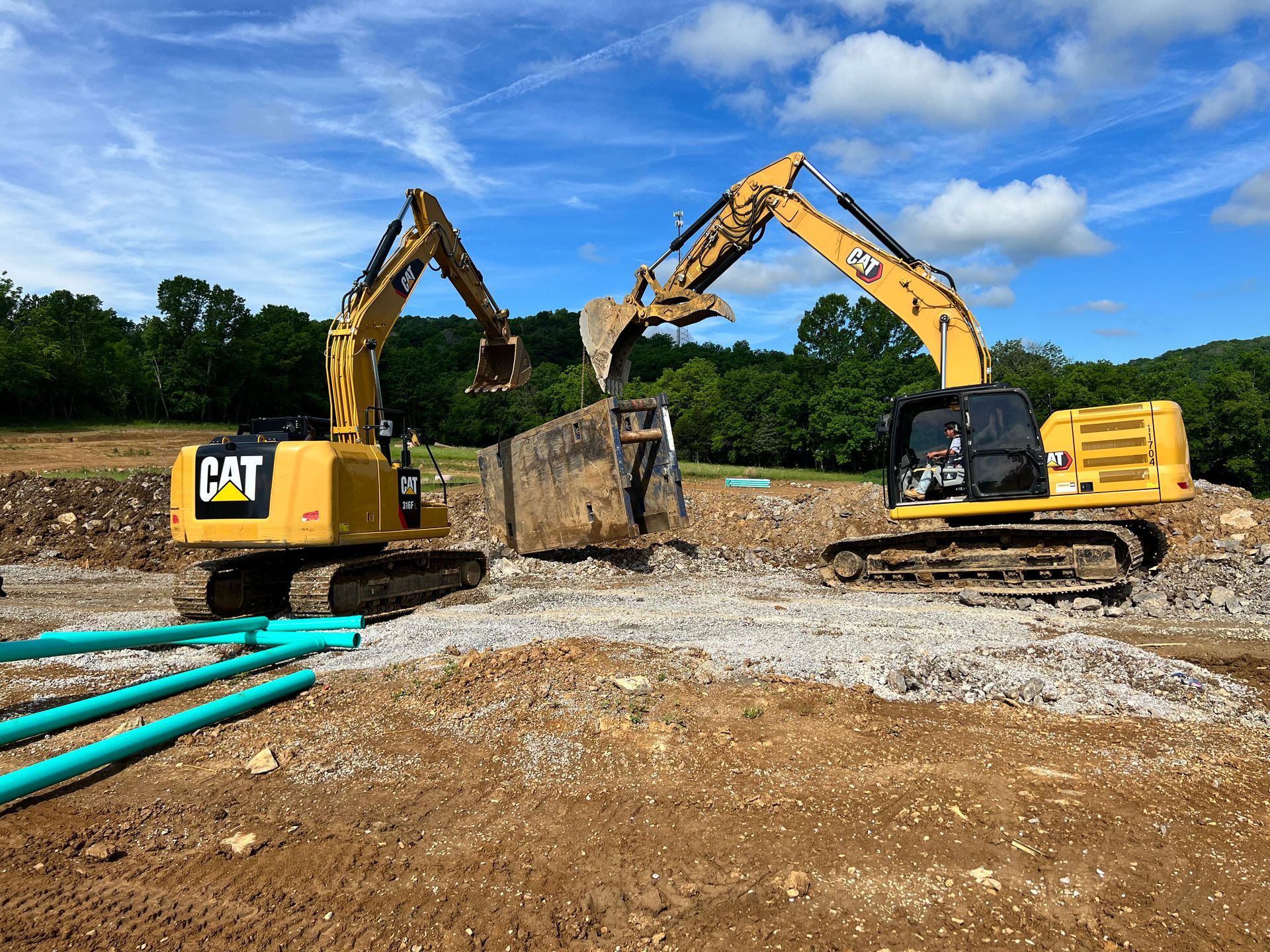 Two yellow CAT excavators moving a metal structure at a construction site. Blue pipes and gravel visible.