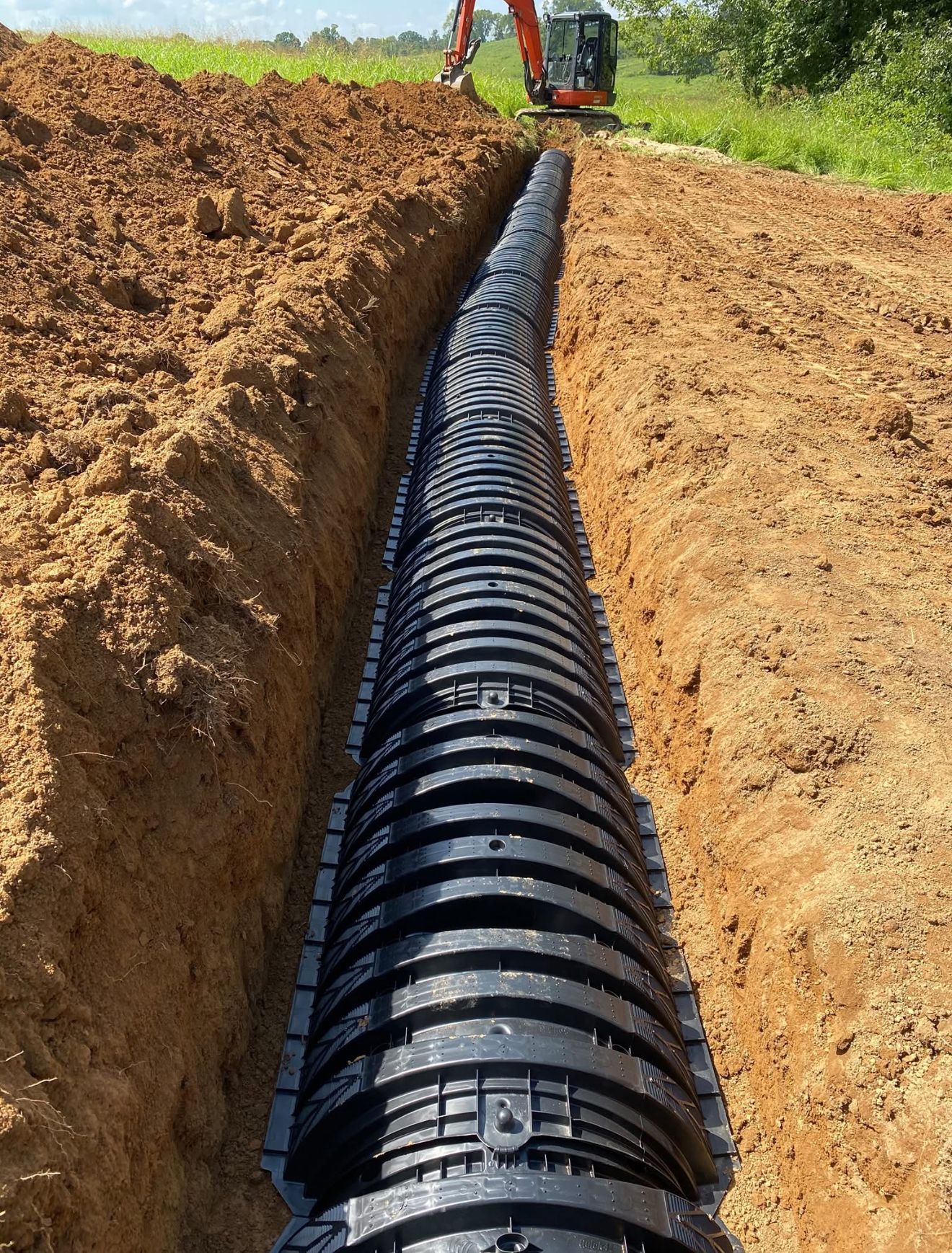 Black drainage pipe installed in a trench, orange excavator in the background.