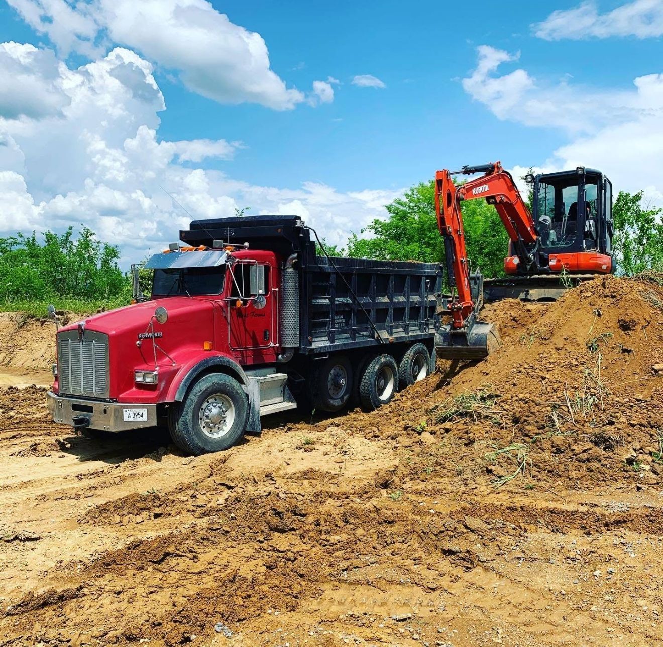 Red dump truck being loaded with dirt by a small excavator under a blue sky.